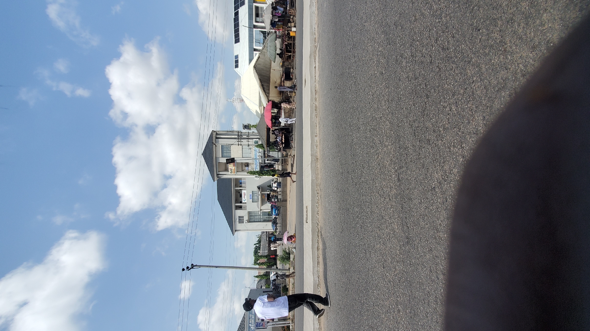 In Jalingo, Nigeria, a sunlit daytime scene unfolds along a paved road with a sidewalk. A man in a white t-shirt and dark pants walks left across the road. On the sidewalk, a few people are visible. A modern, multi-story building with a dark roof stands prominently. To its right, a row of market stalls with awnings and umbrellas are set up, displaying goods. Some individuals are seen interacting at these stalls. The sky is blue with scattered white clouds, and power lines are present. The foreground shows the asphalt of the road and a dark shadow.