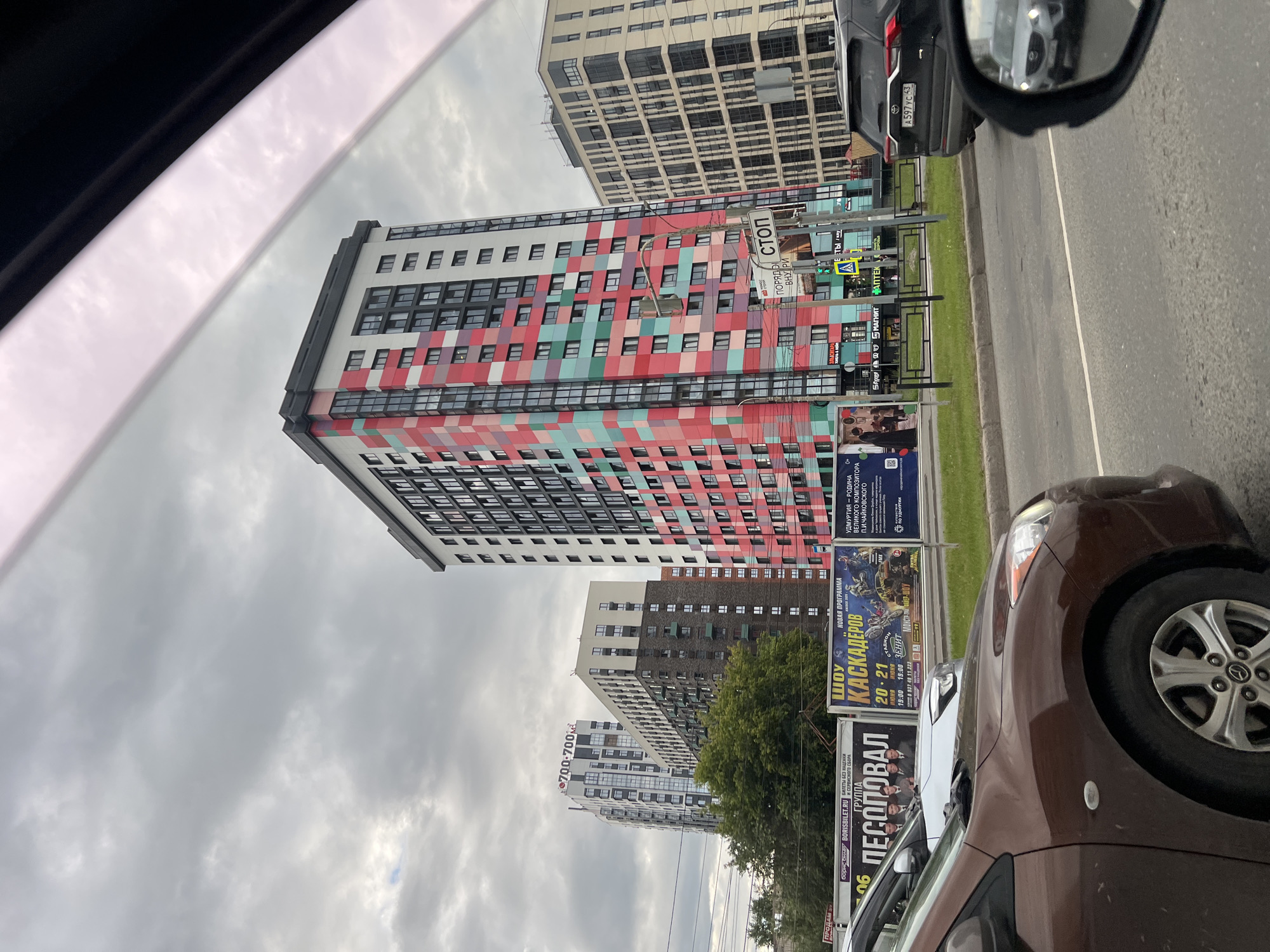 This is a photograph taken from inside a vehicle, looking out at a street scene in Izhevsk, Russia. The camera is angled, making the buildings appear to lean. The sky is overcast and cloudy.
The primary subjects are several multi-story apartment buildings with colorful facades. One building in the foreground features a distinctive pattern of red, pink, and teal rectangles. Another building behind it is lighter in color with a grid of windows. Further down the street, another building is visible.
There are several billboards and signs along the street. One billboard advertises a "SHOU KASKADEROV" (Stunt Show) with dates and times. Another sign mentions "BORISBILET.RU" for ticket sales. There are also signs for "MAGNIT" and "APTEKA" (Pharmacy). A traffic sign that reads "СТОП" (Stop) is visible, as well as a pedestrian crossing sign.
In the foreground, parts of two cars are visible. The closest is a dark brown car, partially obscuring the view of the street. A portion of a black car with a license plate reading "A597 YC 43" is also visible. The license plate format suggests it is Russian.
The time of day appears to be daytime, given the visibility, but the overcast weather makes it difficult to be precise. The overall impression is of a modern urban environment on a cloudy day. The bright colors of the buildings contrast with the muted sky and asphalt of the road.