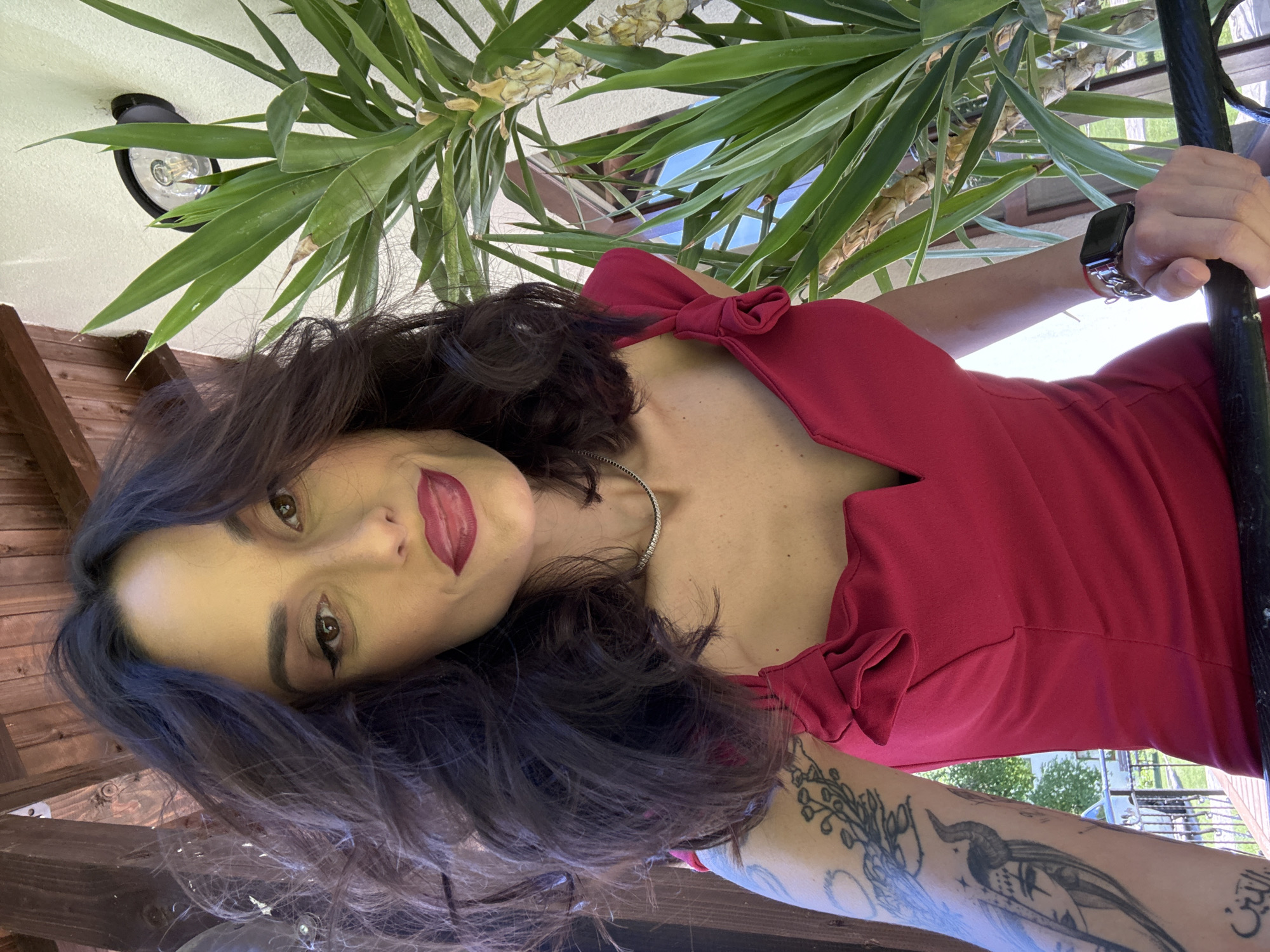 A woman in a red dress is looking at the camera. She has dark hair, fair skin, and red lipstick. She is standing outside under some greenery. She is holding onto a black pole with her right hand, and she is wearing a watch on her wrist. She has tattoos on her right arm. The location is Ribaritsa, Bulgaria. A light fixture is mounted on the wall above her.