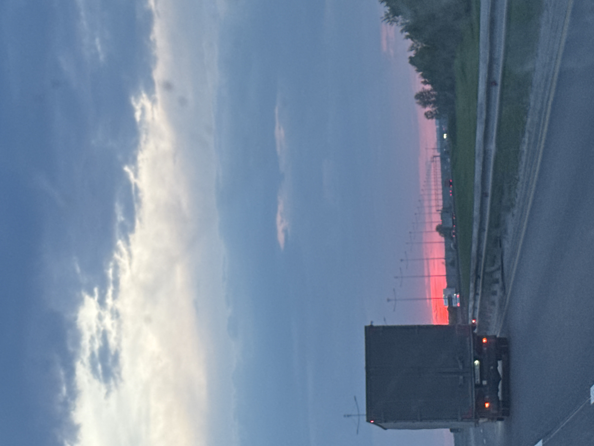 A photograph taken in Кондратово, Russia, shows a large truck on a highway during a sunset. The sky is a mix of blues and pinks with cloud formations. The road runs along the right side of the frame and is lined with a guardrail and trees. Streetlights are visible in the distance. The truck is dark in color and its taillights are illuminated.