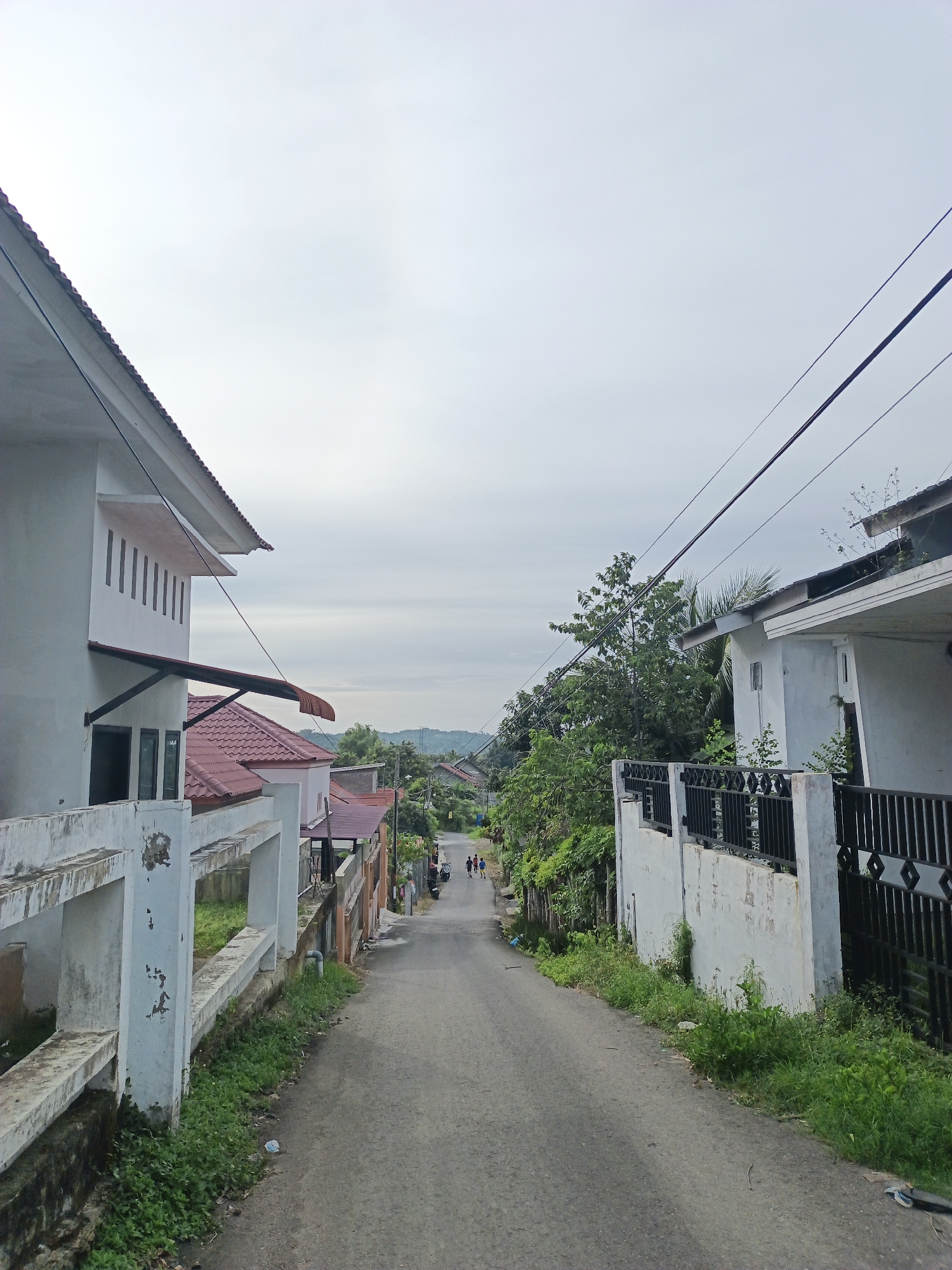 The image shows a paved street in Lhokseumawe, Indonesia, sloping downward into the distance. The sky is overcast. On either side of the street are houses with white walls and fences. The house on the left has a red tile roof and a cantilevered awning. The house on the right has a black metal fence. Power lines run along the street above the houses. In the distance, several people are walking down the street. Green vegetation grows along the edges of the road and between the houses.