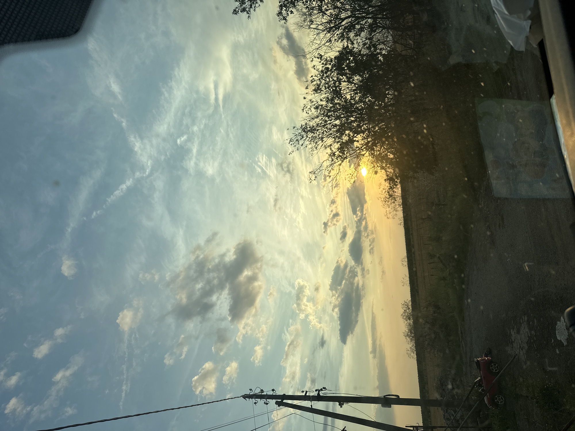 The image depicts a rural sunset scene in Mlekarevo, Bulgaria. A power line pole stands to the left, with visible wires and insulators. In the foreground, a small red toy car is parked on a gravel path. Beyond the path, a field stretches towards the horizon, bordered by trees and bushes. The sky is filled with clouds of varying shades of blue, gray, and yellow, illuminated by the setting sun, which is partially obscured by foliage in the mid-right of the frame. The overall impression is one of natural beauty and a serene, quiet atmosphere.