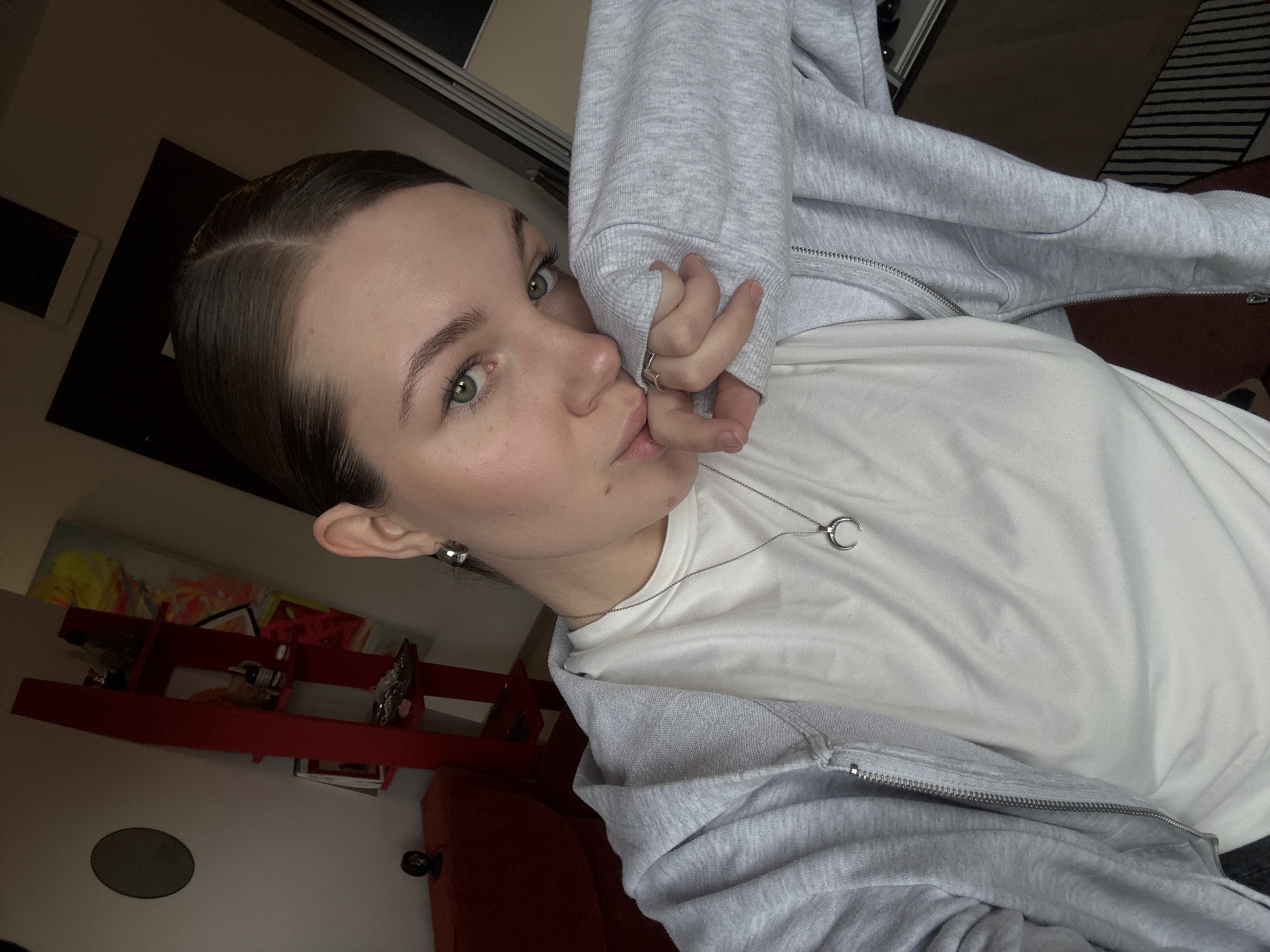 An indoor selfie captures a female subject positioned in a residential setting within Vozdovac Urban Municipality, Serbia. The subject, of fair complexion, has dark brown hair parted centrally and pulled back. Her eyes are green. She wears a light grey zippered hoodie over a white crew-neck t-shirt. Silver hoop earrings are visible on her ears, and a silver crescent moon pendant hangs from her neck. A silver ring is present on her right ring finger. Her right arm is bent, with the hoodie sleeve near her mouth, and her right hand partially obscures her lower face as she looks directly at the camera.
The background on the left features a white wall with a black circular clock and a partially visible red sofa or armchair. A small black figurine rests on the furniture or an adjacent surface. A tall, narrow, red shelving unit with multiple tiers displays small objects, including a bottle and items in a tray. Behind the shelving unit, a vibrant abstract painting with yellow, pink, and blue hues is partially visible. The right side of the background reveals a dark vertical panel, possibly a door or wardrobe, and a section of a floor covered by a striped rug featuring black and light-colored bands. Lighting appears to be even and diffused.