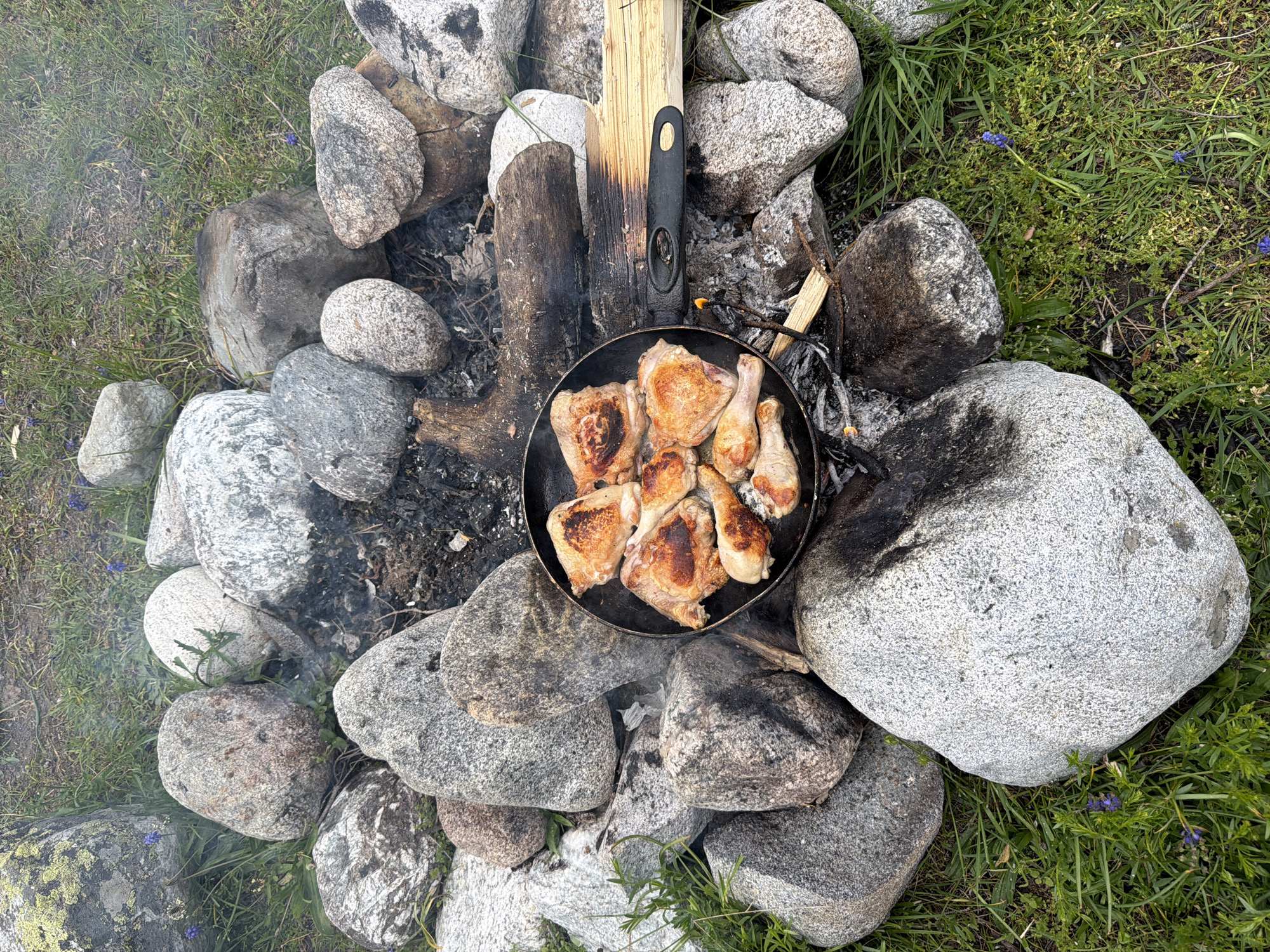 The image depicts an outdoor cooking scene amidst a rocky, grassy environment, possibly in the vicinity of Mazeri, Georgia. A black frying pan filled with pieces of cooked chicken is placed on a makeshift campfire constructed from large stones and logs. Smoke rises from the fire, indicating it is still active. The chicken pieces appear browned and well-cooked. The surrounding area is covered in green grass and scattered small purple flowers. The rocks are of various sizes and textures, some appearing to be granite. The overall impression is one of rustic, outdoor food preparation. No people are visible in the frame.