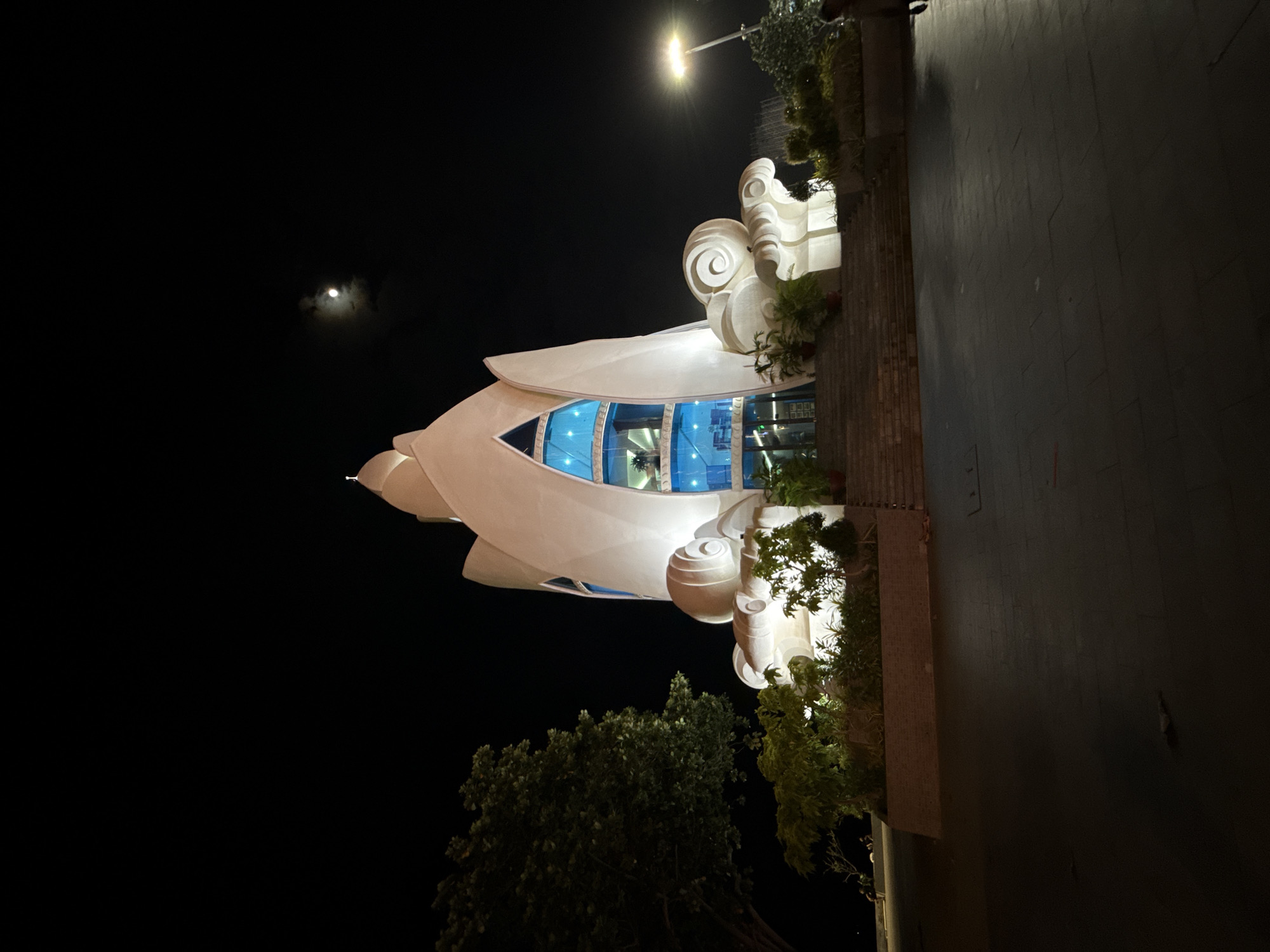 The image shows the Nha Trang Center, a shopping mall in Nha Trang, Vietnam, at night. The building's facade is illuminated, and its distinctive lotus-shaped design is visible. There is a full moon in the dark sky, with some clouds. The foreground is a paved area, and there are some trees and plants surrounding the building. A street lamp is visible on the right side of the image. There are a few people in the distance, but they are not clearly identifiable. The overall scene is one of a modern architectural landmark at night.