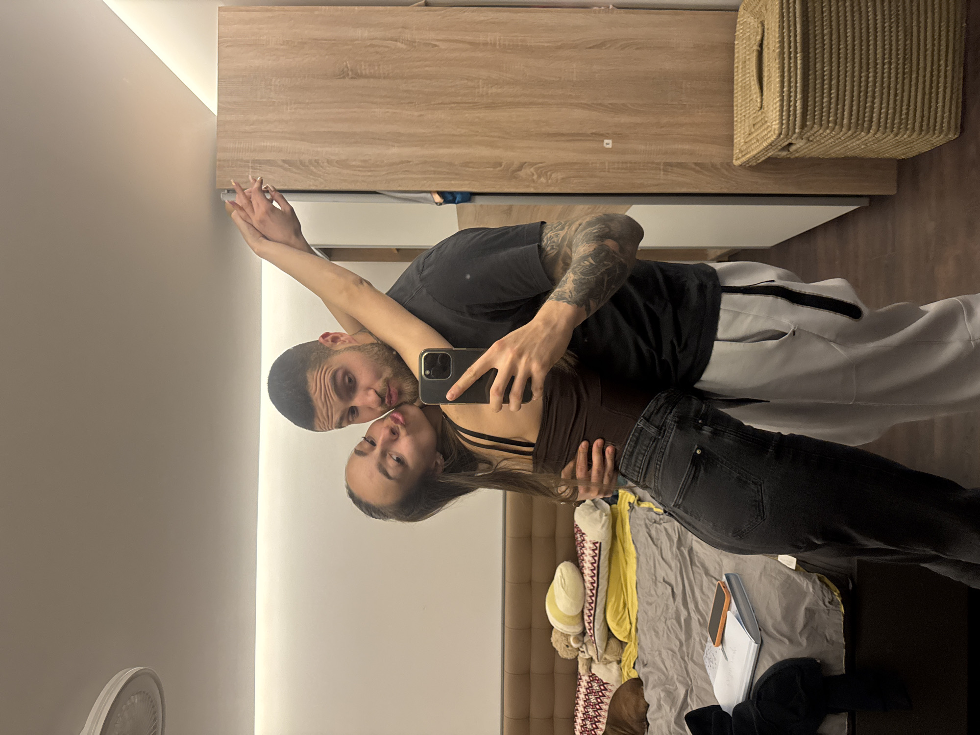 A man and a woman are taking a selfie in a mirror. The man is wearing a black t-shirt, grey sweatpants, and has tattoos on his left arm. The woman is wearing a brown top and black jeans. They are standing in a bedroom, with a bed and furniture visible in the background. The location is Sofia, Bulgaria.