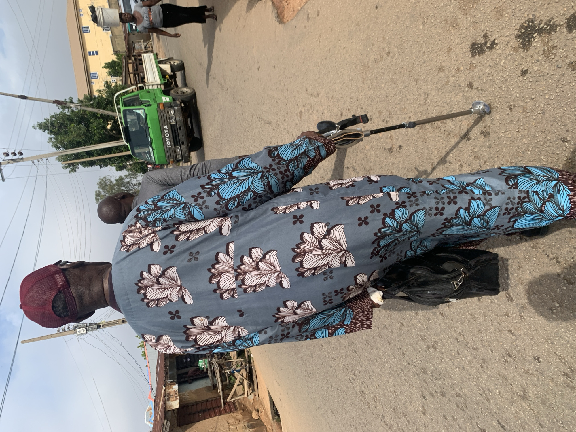 In Jos, Nigeria, an elderly man with dark skin, wearing a patterned blue and white traditional outfit and a maroon baseball cap, walks down a paved street. He is using a metal cane for support and carries a black bag. He is accompanied by another man, partially visible, also walking. A green Toyota truck is parked on the side of the road, and a woman with a bucket on her head walks in the distance. The street is lined with buildings and utility poles with visible wires. The sky is cloudy.