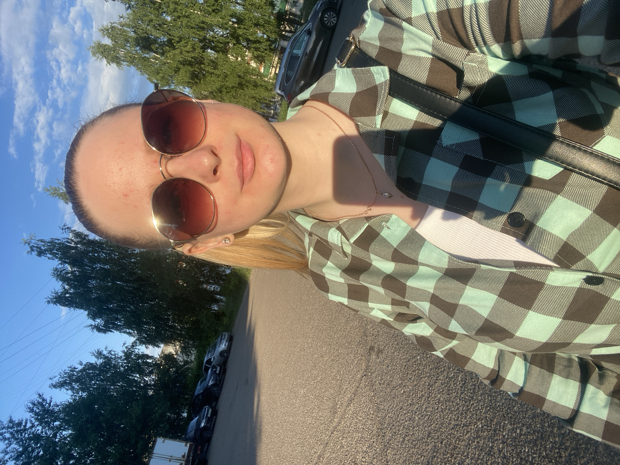 A young woman is taking a selfie outdoors in what appears to be a parking lot. She is wearing a plaid shirt over a white top, large sunglasses, and a necklace. A black bag is slung across her shoulder. In the background, several cars are parked, and trees line the edge of the lot. The sky is blue with scattered clouds, and the sun is casting long shadows, indicating it is late afternoon or early evening.