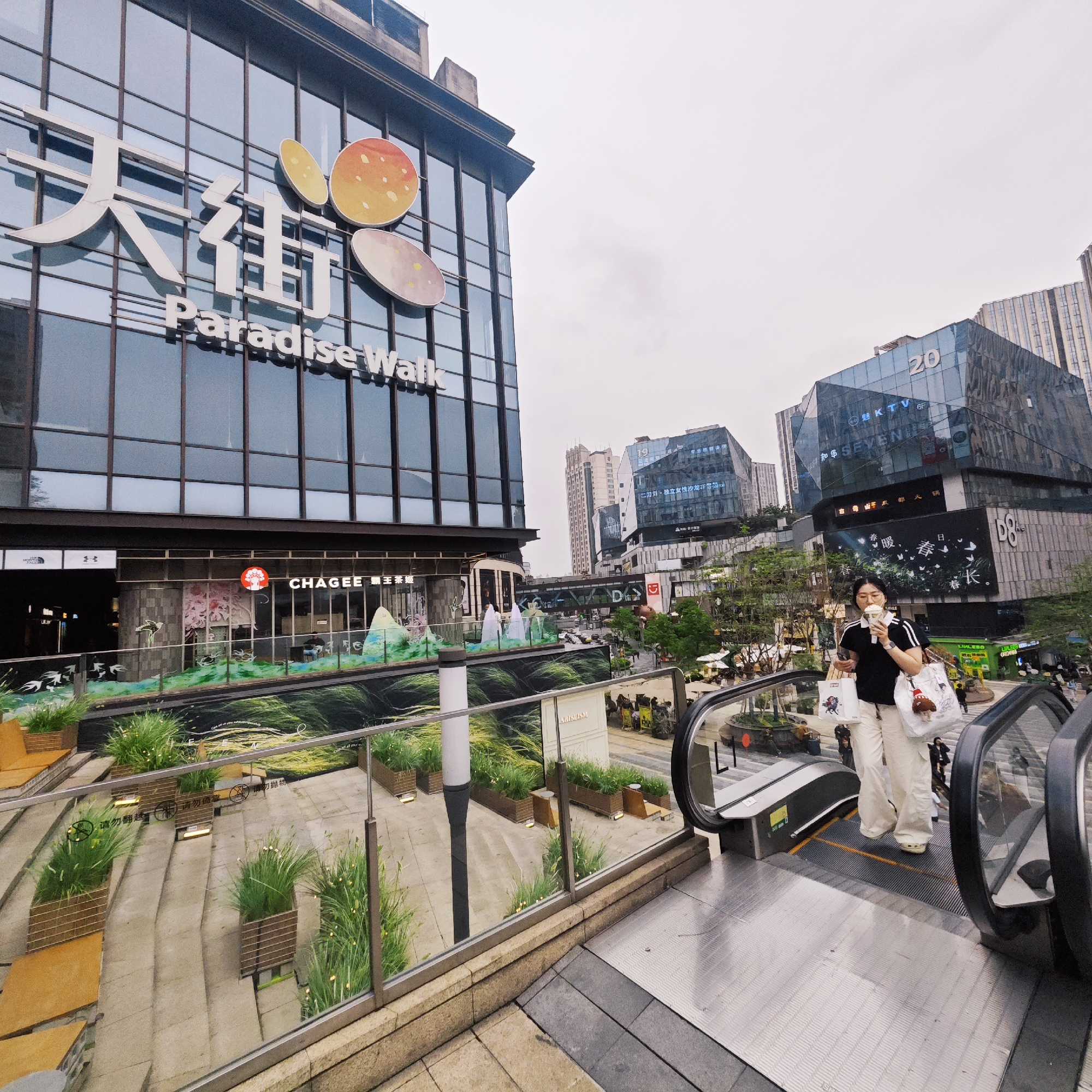 The image captures a street scene in the Yuzhong District of Chongqing, China. A large glass building bearing the name "Paradise Walk" with accompanying Chinese characters is visible on the left. Below the signage is a CHAGEE store. The street scene includes a landscaped area and an escalator. On the escalator, a person wearing a black shirt and white pants is ascending, holding a beverage and shopping bags. In the background, various buildings with signs are visible, including one with the number "20." The weather appears overcast.