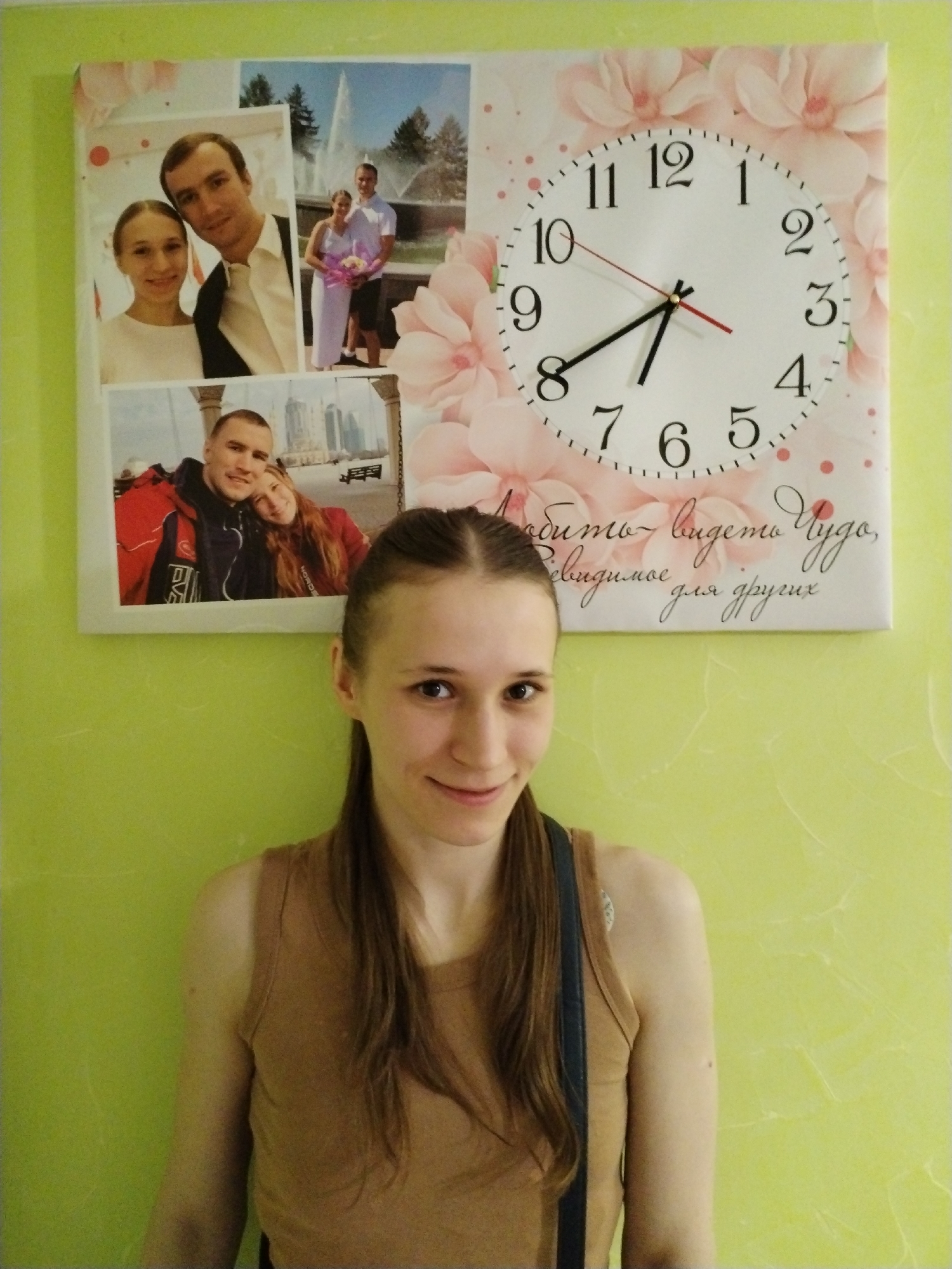 A young woman with long, straight brown hair pulled back in a ponytail stands in an indoor setting, smiling directly at the camera. She wears a light brown tank top, with a dark strap visible over her left shoulder. The background is a textured light green wall.
Mounted on the wall behind her is a large, rectangular canvas print that serves as a clock and a photo display. The clock face, positioned on the right side, is white with black numerals (1-12) and shows the time as approximately 8:40 with black hour and minute hands and a red second hand. The clock face is set against a decorative background of soft pink floral patterns, possibly cherry blossoms, and small pink dots.
The left side of the canvas features three framed photographs of a couple, likely the woman's family or partner, integrated into the design. The top-left photo shows a smiling couple in a close-up. The top-right photo depicts the same couple outdoors, standing near a fountain with trees, where the woman is holding a bouquet, suggesting a special occasion such as a wedding or engagement. The bottom-left photo shows the couple in a different outdoor setting, possibly a city, with the man in a red and black jacket and the woman leaning on him.
Below the clock and photos, elegant Russian script is visible on the canvas. The text reads: "Любить – видеть чудо, невидимое для других," which translates to "To love is to see a miracle, invisible to others."
The scene takes place in what appears to be a home in Chaikovsky, Russia, characterized by personalized decor reflecting sentimental memories. The woman's pleasant expression and the personal nature of the wall art convey a warm and inviting atmosphere.