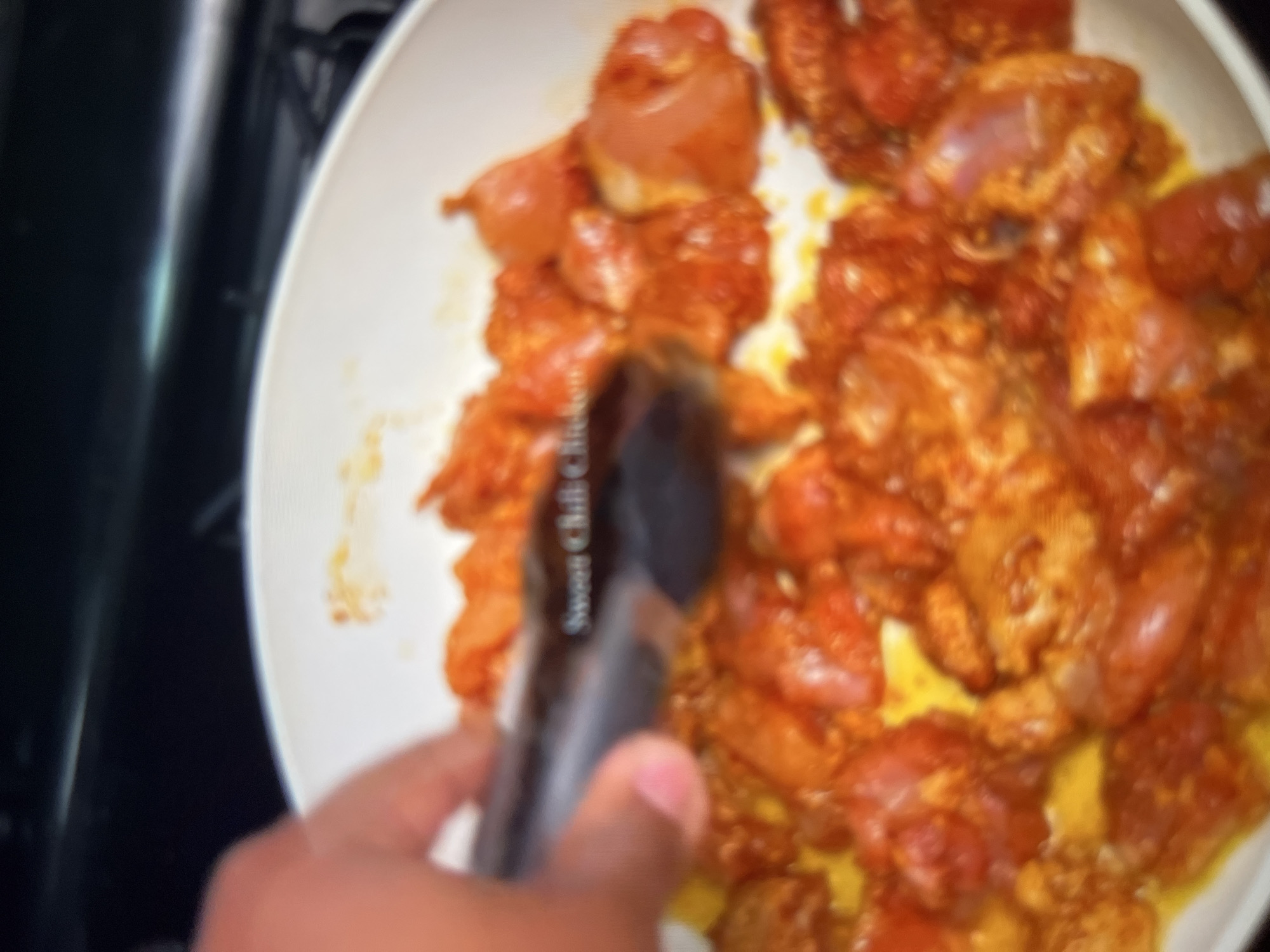 A person's hand uses black tongs to flip pieces of raw chicken coated in a reddish-orange spice blend in a white pan. Some of the chicken pieces are cut into bite-sized chunks, while others are larger. The pan appears to be on a stovetop. A label on the tongs reads "Sweet Chili Chicken." The background is dark and out of focus. There is no visible indication of location in Abuja, Nigeria.