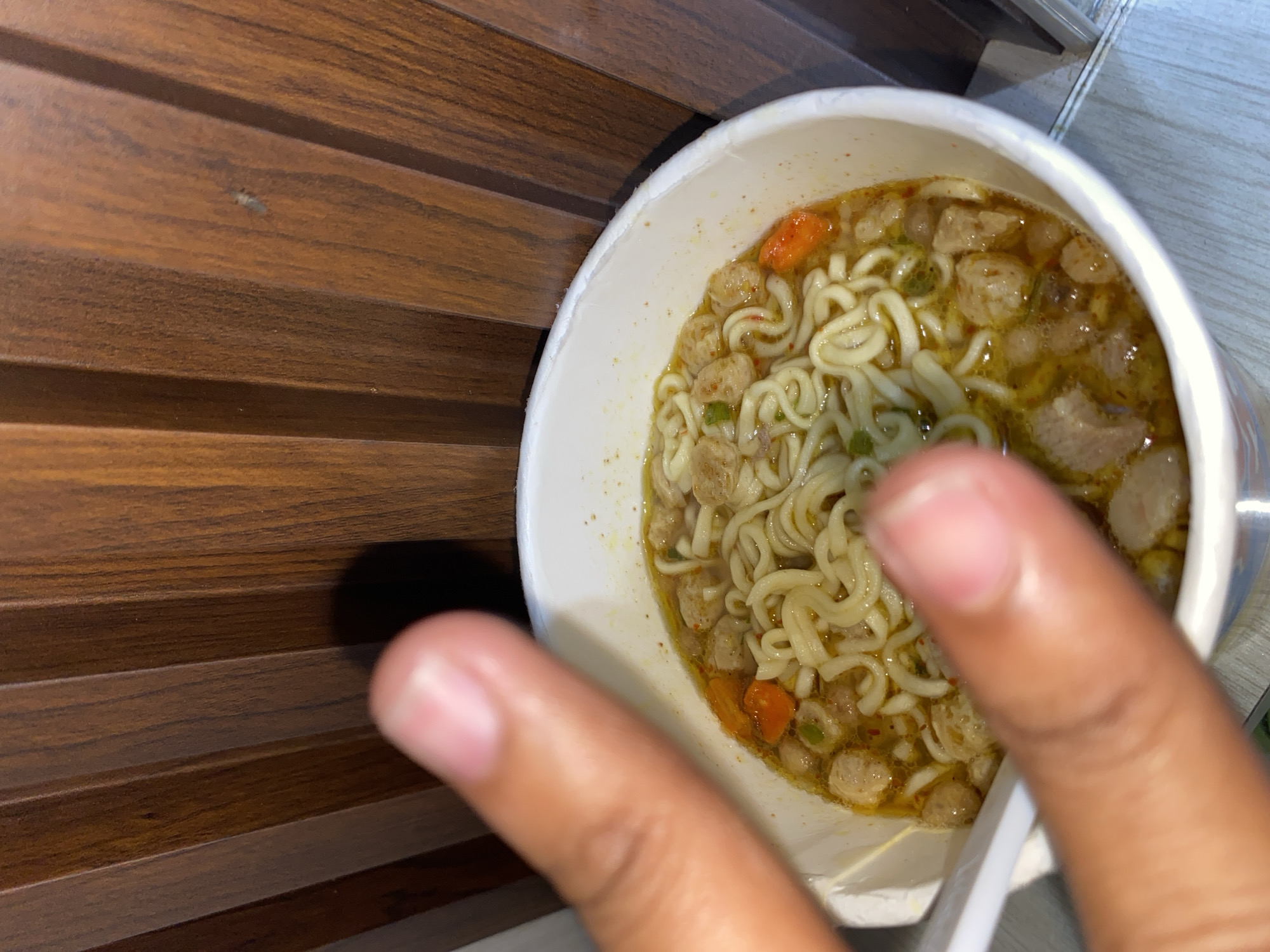 A close-up shot shows a cup of instant noodles, likely ramen, filled with broth, noodles, meatballs, and small pieces of vegetables. A hand is partially obscuring the view of the noodles, with two fingers visible in the foreground. A white plastic fork is partially visible inside the cup. The background features a wooden surface on the left and a light-colored tiled surface on the right. The location in the city of Lhokseumawe, Indonesia, is not directly determinable from the image content, although it is implied based on the prompt.