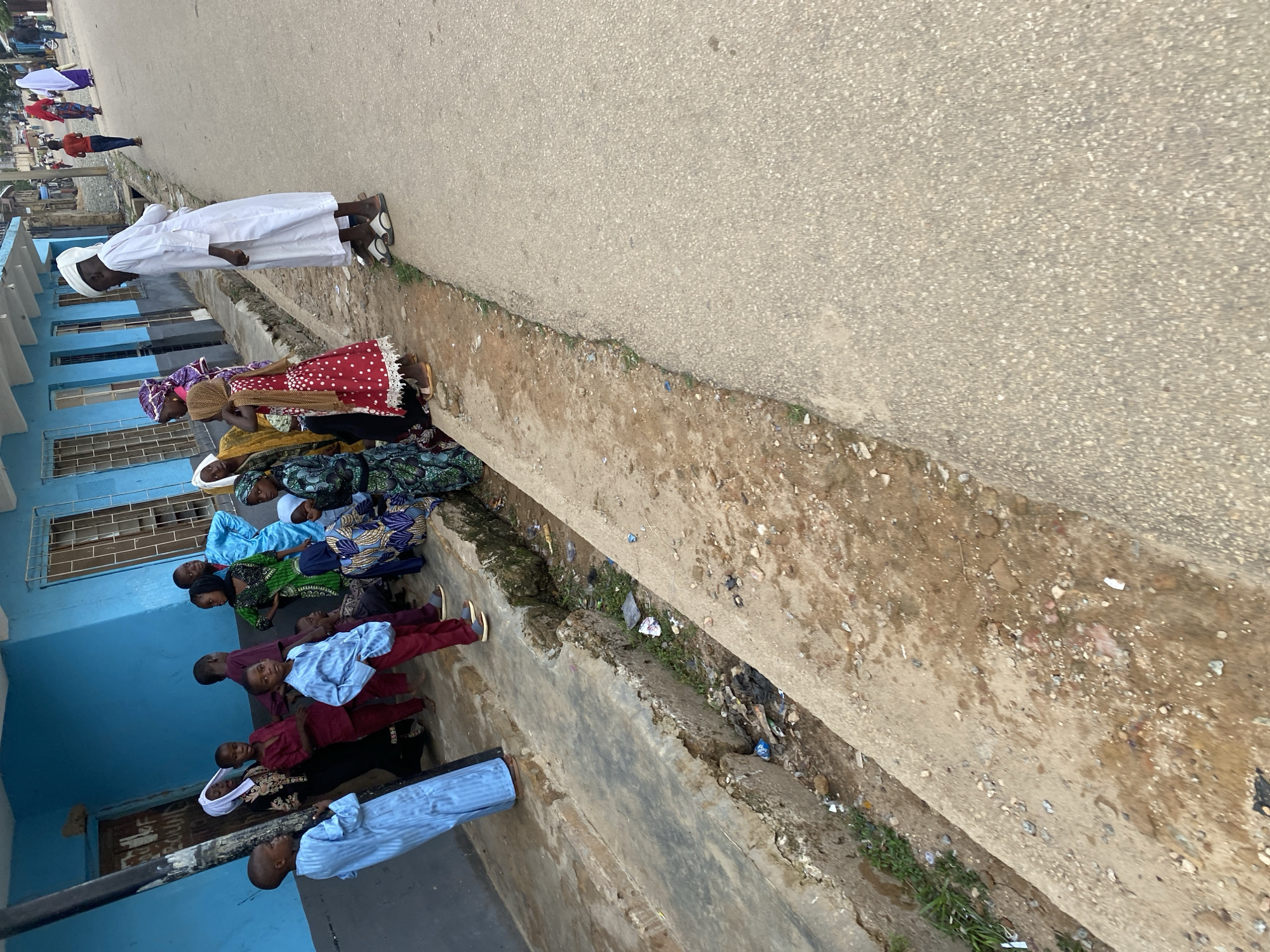 A group of people, predominantly children, are gathered on a dirt and concrete pathway next to a blue building. The building has windows with bars and appears to be a commercial or residential structure. Some of the children are wearing traditional clothing, including headscarves and long robes. One man, dressed in a white robe and head covering, stands on the asphalt road adjacent to the pathway. A concrete drainage ditch runs along the side of the pathway, containing debris and some vegetation. In the background, other people are visible walking along the road, and the environment suggests a moderately urban setting with other buildings and trees. The scene captures a moment of everyday life, possibly in a neighborhood in Jos, Nigeria, given the context.