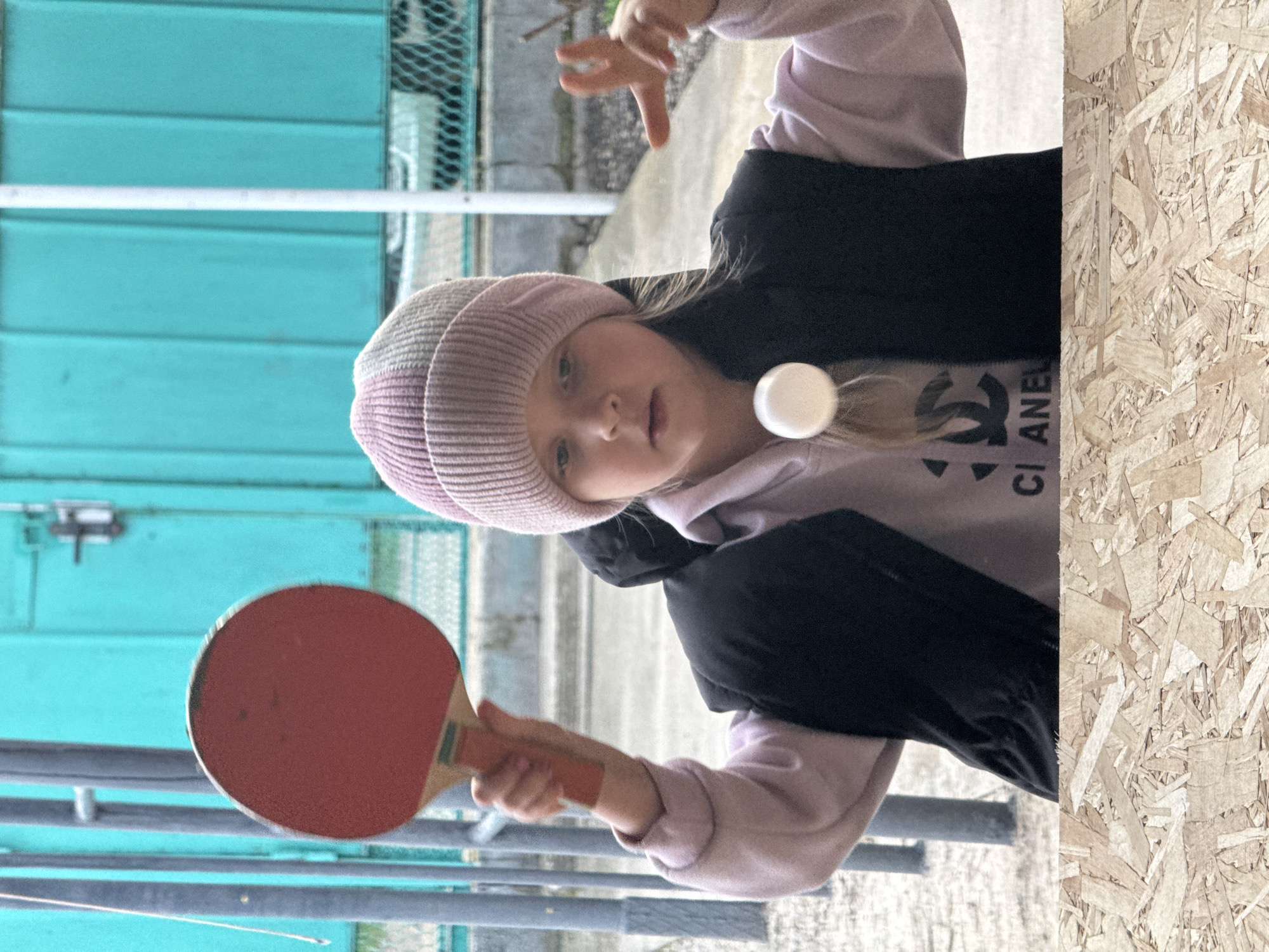 A young girl in Besskorbnaya, Russia is playing ping pong outdoors. She is wearing a pink and gray striped beanie, a light pink hoodie with a black Chanel logo, and a black puffy vest. She holds a red ping pong paddle in her right hand and has a white ping pong ball floating in the air near her face. To the left of the girl is a blue wall with horizontal slats and metal bars. To the right is a wall made of particle board.