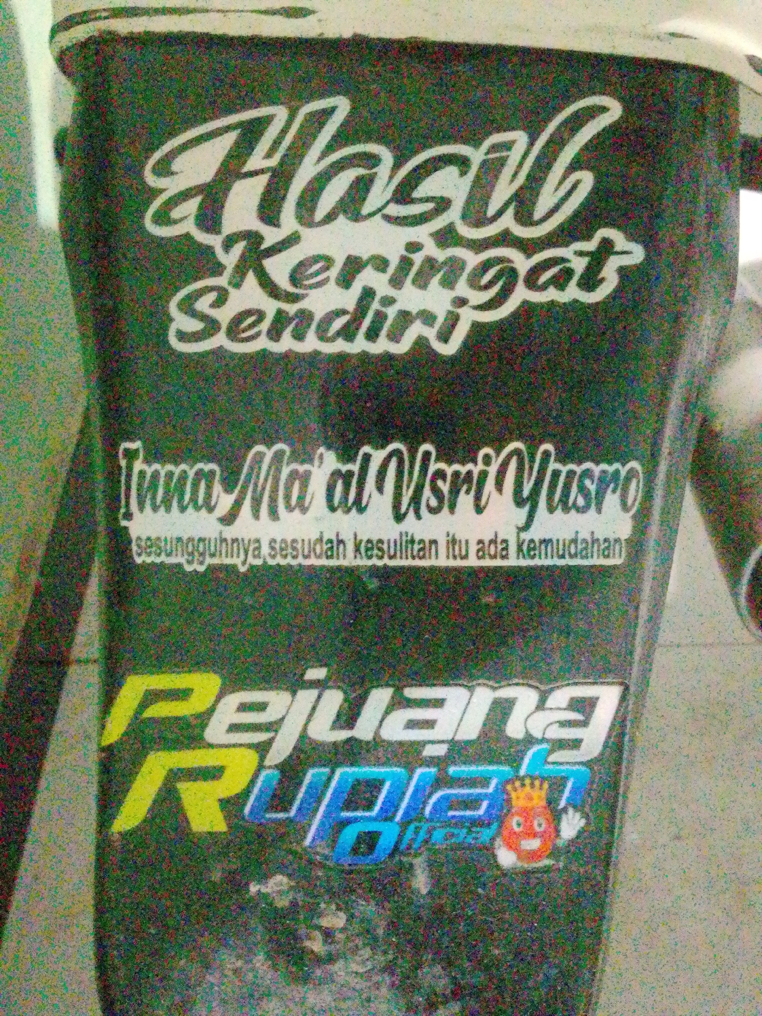 The image displays a close-up of a dark-colored object, possibly a vehicle or equipment, adorned with text and a cartoon character sticker. The text is in white and features Indonesian phrases. The top portion reads "Hasil Keringat Sendiri" (Result of Own Sweat). Below this, in a stylized font, is "Inna Ma'al Yusro" followed by smaller text that reads "sesungguhnya sesudah kesulitan itu ada kemudahan" (Indeed, after hardship there is ease). The bottom features larger, colorful text: "Pejuang Rupiah" (Rupiah Fighter), with a small, cartoonish character of a red, crowned figure with a smiling face and a raised hand on the right side of the text. The background is blurred and appears to be a textured surface, possibly concrete or a wall. The overall impression is that of decals or stickers applied to a surface. The image itself has a grainy texture, suggesting it was taken in less than ideal lighting conditions. There is no indication of the city of Bandar Lampung in the image itself; this information is external to the visual content.
