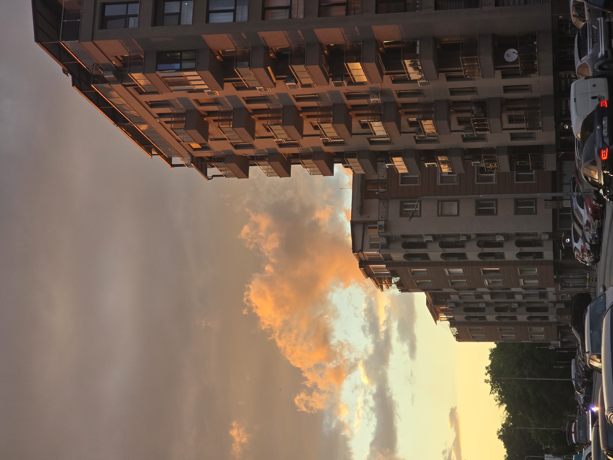 Captured from a ground-level perspective, the scene features two multi-story buildings positioned on the left and right sides of the frame. The buildings' architectural details include rows of windows and balconies. A parking area is visible on the right side of the frame, occupied by several parked vehicles. In the center, the sky transitions from a gradient of warm yellow and orange hues at the bottom to darker, greyish tones at the top. Some clouds can be seen in the sky. The location is Unknown, Unknown.