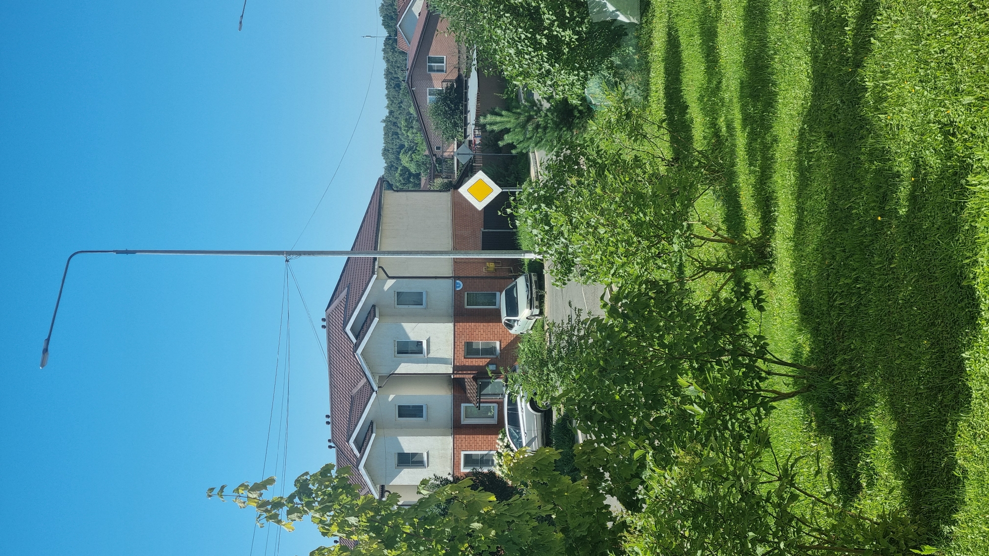 A sunny day with a clear blue sky is depicted in this image, likely taken in the city of Верзилово, Russia, given the context. The foreground is dominated by lush green grass and trees, with shadows cast by the foliage. In the midground, a street is visible with two cars parked. One is a silver SUV, and the other is a silver sedan. Behind the street, a modern two-story duplex-style house with a brown roof and cream-colored walls stands prominently. The house has multiple windows and a brick lower level. A yellow diamond-shaped road sign is positioned to the right of the house. In the background, more houses and a line of trees can be seen against the horizon. A tall street lamp is also visible on the left side of the image.
