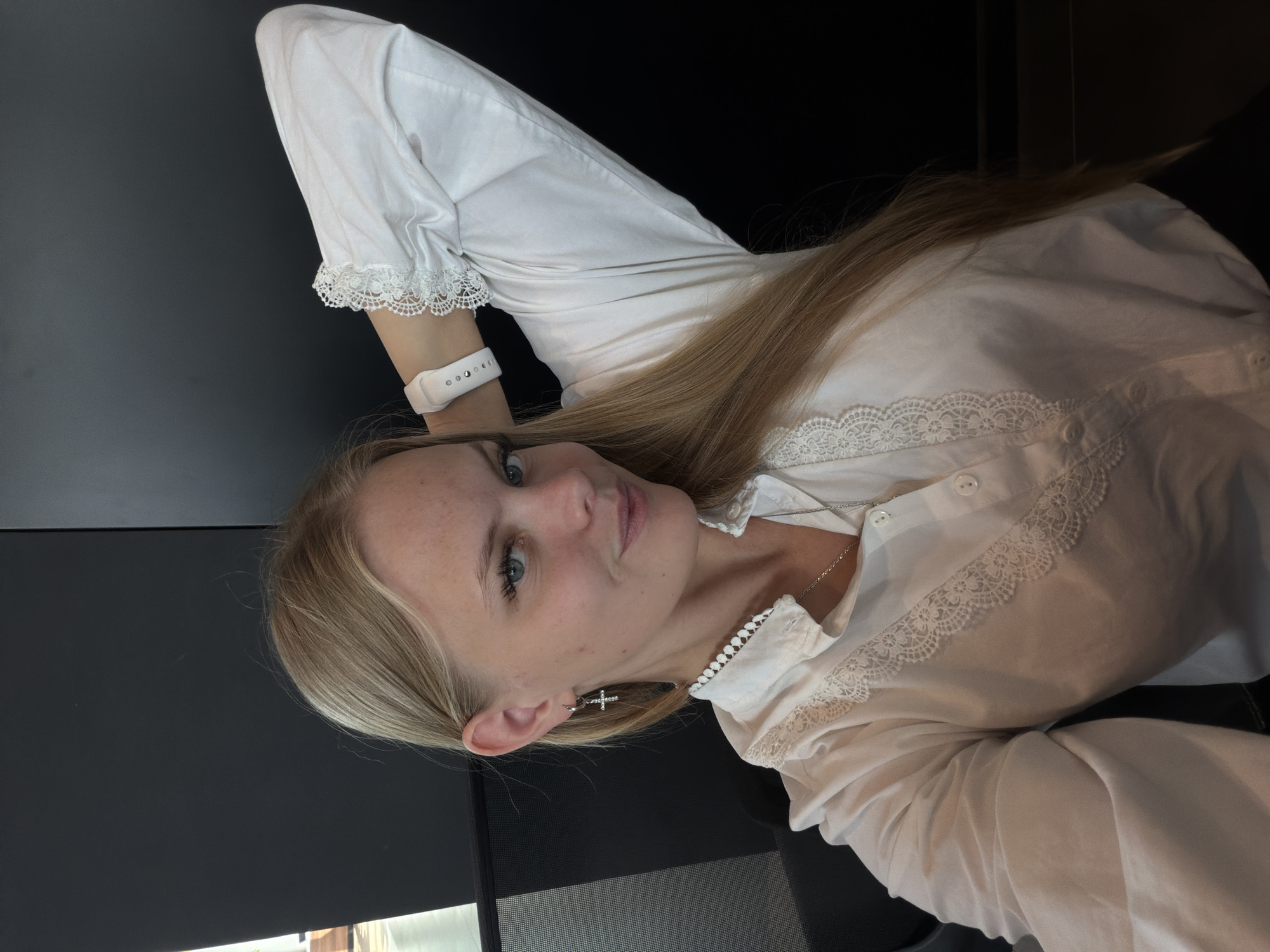A young woman with blonde hair is captured in a selfie, sitting in an office setting with a dark wall behind her. She is wearing a white blouse with lace trim around the collar and cuffs, and a silver cross earring in her left ear. A white smartwatch is visible on her left wrist. Her right arm is behind her head, and she is looking at the camera with a slight smile. The image does not provide enough information to determine the specific location within Izhevsk, Russia, or any other details about the environment.