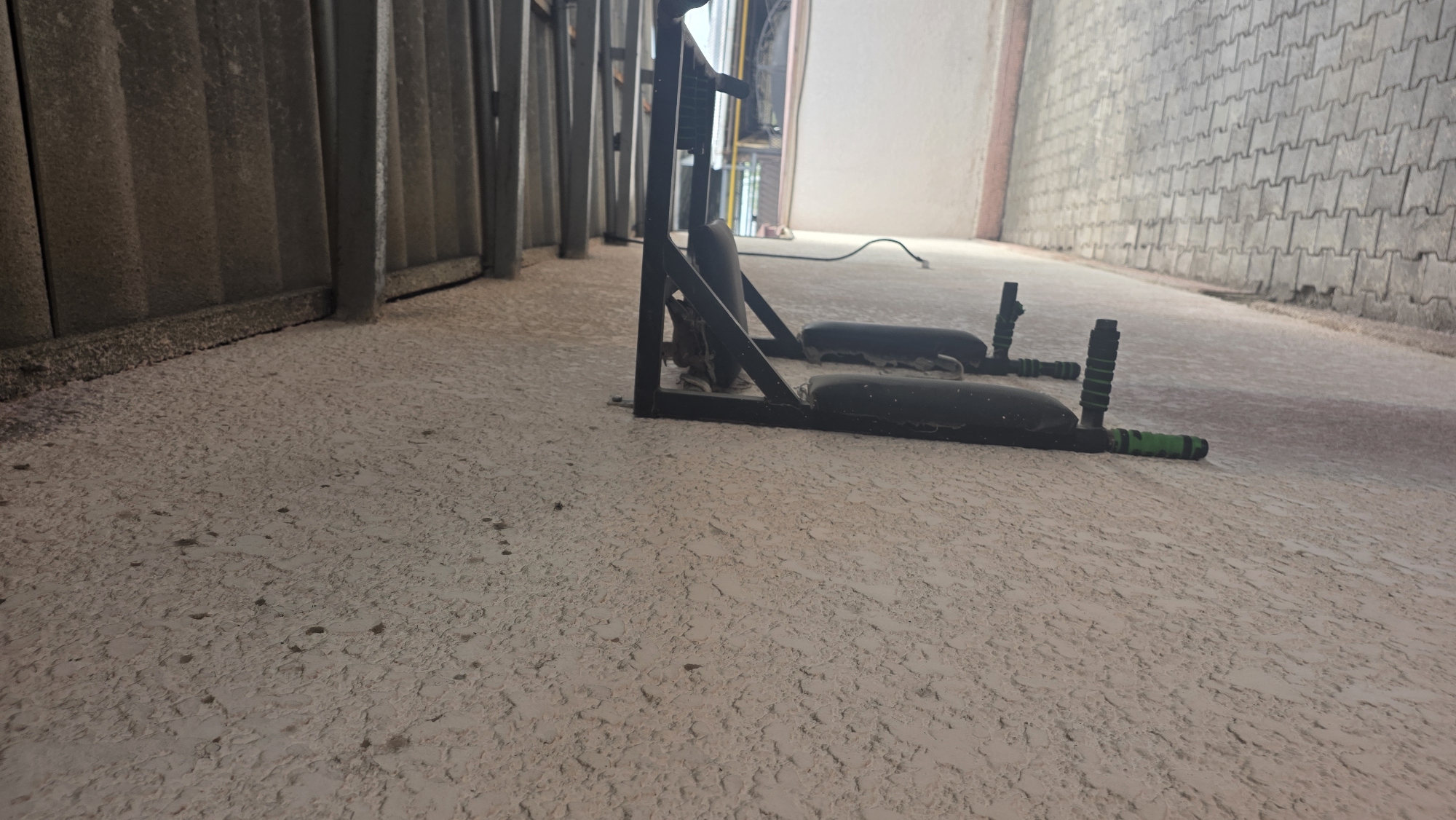 This image depicts an exterior view of a building in Chuy, Kyrgyzstan. The focus is on a wall with a textured, light-colored finish, possibly stucco or concrete, showing some signs of wear and dirt. Mounted on this wall is a black metal apparatus with two dark, cylindrical objects hanging from it. These objects appear to be some form of weights or training equipment, with green handles visible on one of them. The apparatus is attached to the wall with brackets. Above the wall, a corrugated metal roof structure is visible, supported by metal beams. The ground in front of the wall is paved with interlocking gray pavers. In the background, a light-colored wall and some yellow pipes are discernible, suggesting an urban or industrial setting. The lighting suggests it is daytime, possibly overcast. There are no people visible in the image.