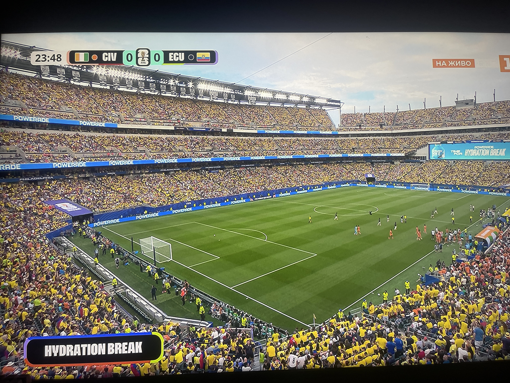 The image shows a wide shot of a soccer stadium packed with spectators. The score graphic at the top indicates the game is between CIV and ECU, with the score tied at 0-0 in the 23rd minute. The majority of the fans are wearing yellow, suggesting they are supporting one of the teams. A banner across the field announces "HYDRATION BREAK." The stadium appears to be a large, modern facility with multiple tiers of seating. The sky is overcast. The specific location in the city of Ruse, Bulgaria is not discernible from the provided image or crops.