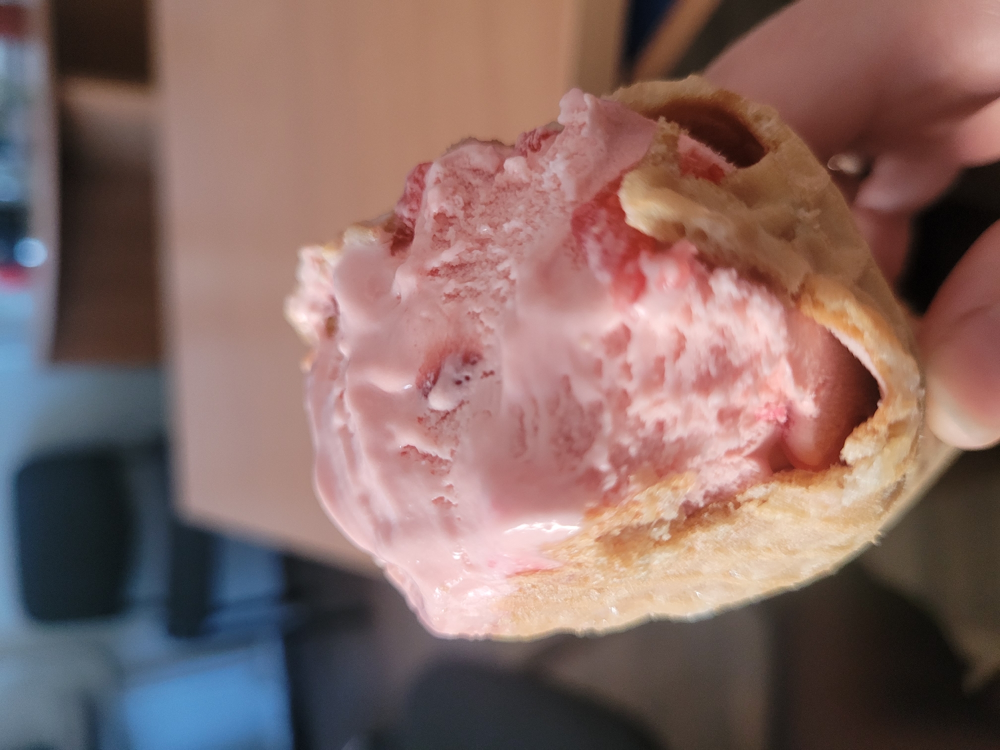 A close-up shot depicts a partially eaten waffle cone filled with pink ice cream, held in someone's hand. The ice cream is textured and appears to be melting slightly. The waffle cone has a visible waffle pattern. The background is blurry, showing parts of a brown box and a hint of a window. The scene does not provide information about the location in Izhevsk, Russia.