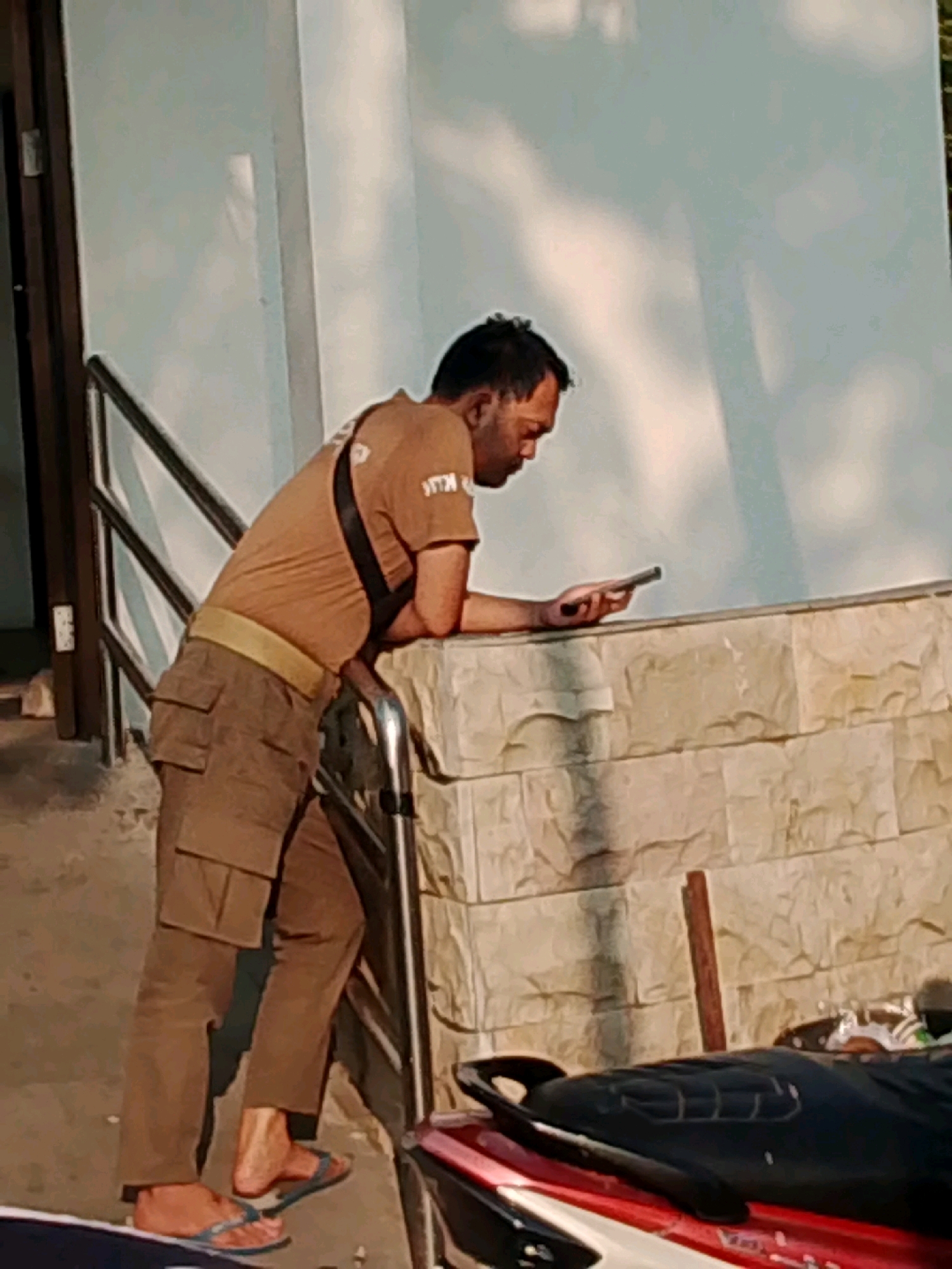 A man is standing near a wall, leaning on a railing and looking at his phone. He is wearing a brown t-shirt with a black strap over his shoulder and brown cargo pants. He has on flip-flops. In the foreground, the seat of a red and black motorcycle is visible. The wall behind the man is made of light-colored stone blocks. The scene appears to be outdoors, possibly in a public area within Probolinggo, Indonesia.