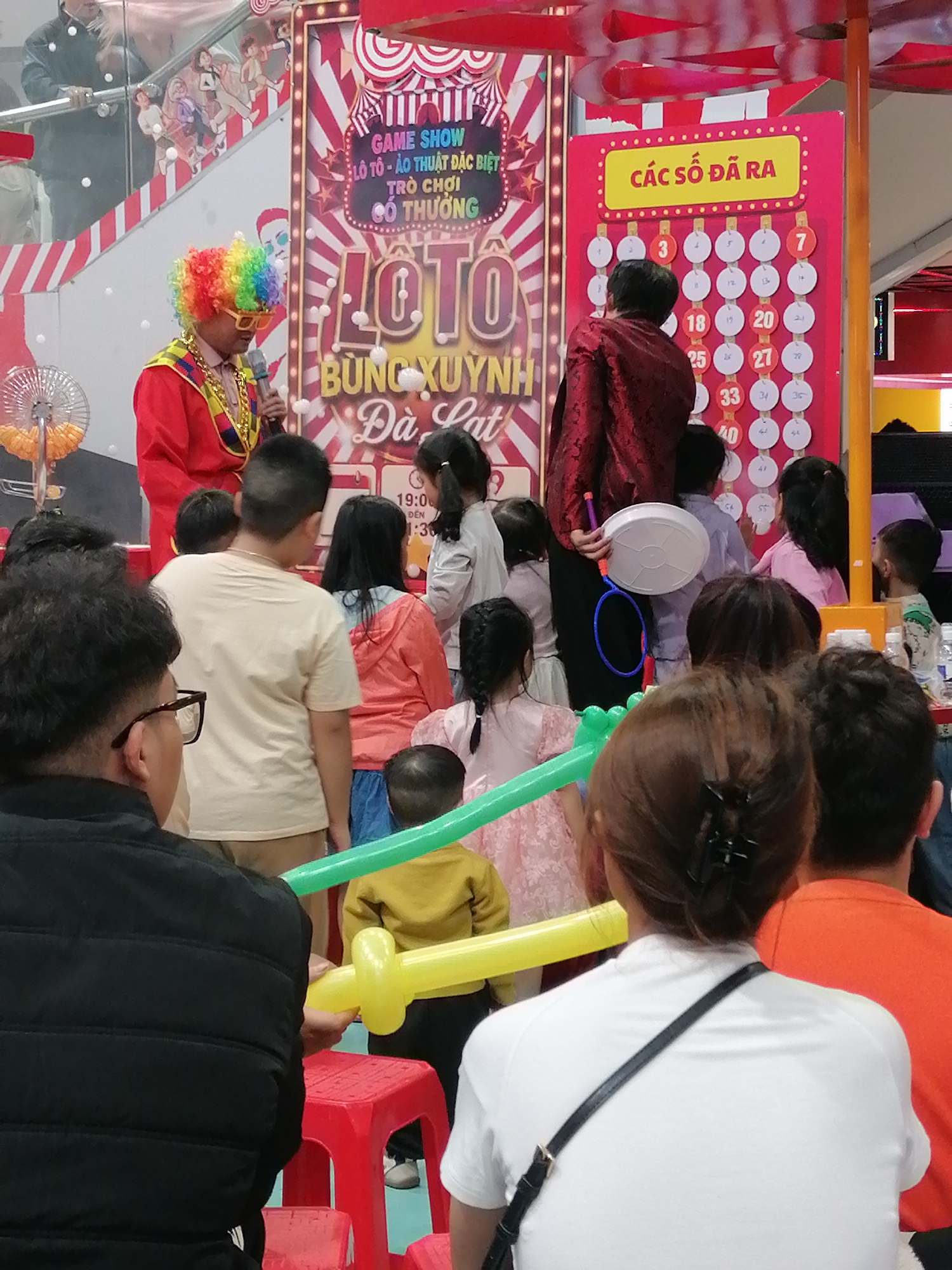 This is a medium shot of a crowded scene in Da Lat, Vietnam, likely at a public event or attraction. In the foreground, a person in a black puffer vest is holding a red stool and a yellow balloon animal. To their right, a woman in a white t-shirt with a black crossbody bag is looking away from the camera. Behind them, a child in an orange shirt is visible, and to the left, a boy in a light-colored t-shirt is looking towards the center of the scene.
The background is dominated by a colorful game show backdrop. A person dressed as a clown with a rainbow wig and yellow glasses is holding a microphone and standing on the left side of the backdrop. The backdrop features large text in Vietnamese, including "GAME SHOW," "LÔ TÔ - ÂU THUẬT ĐẶC BIỆT," and "TRÒ CHƠI CÓ THƯỞNG." Below this, a large graphic with the word "LOTO" is prominently displayed, along with smaller text that appears to be "BUNG XUYNH". To the right of the clown, a man in a red jacket with a patterned design is standing in front of a grid of numbered circles, presumably for a lottery or bingo game. Several of these circles have numbers filled in.
The environment appears to be indoors, possibly a shopping mall or entertainment venue, with a bright, festive atmosphere suggested by the colorful decorations and the presence of a game show. The lighting is bright, indicating artificial illumination. Several other people, mostly children, are visible in the middle ground, observing the events.