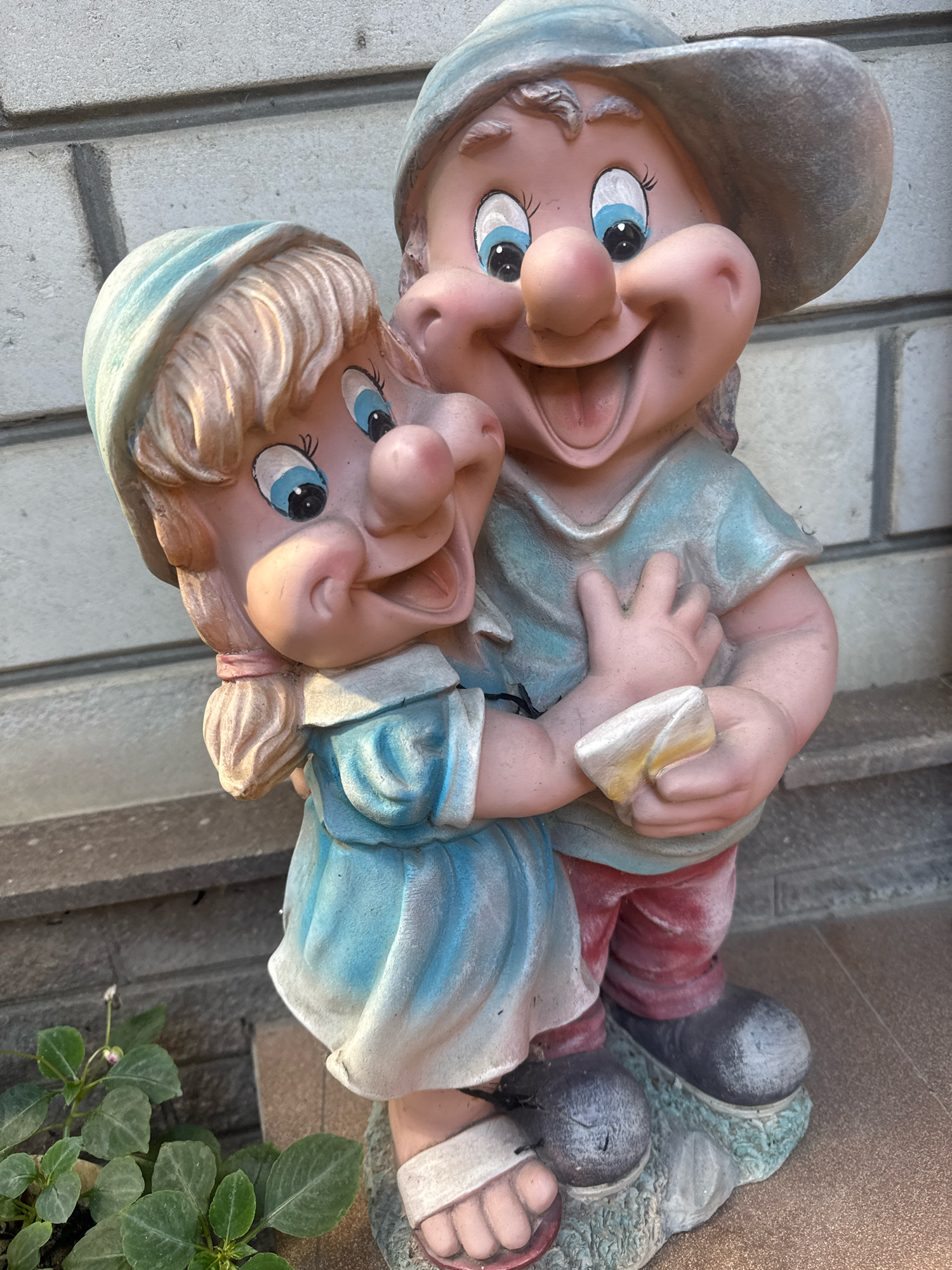 A statue of two gnomes, a male and female, are positioned in front of a brick wall. The gnomes are depicted in a close embrace. The male gnome is on the right, wearing a wide-brimmed hat and a light blue shirt with red pants. He has a large, wide smile and his arms are around the female gnome. The female gnome is on the left, wearing a light blue bonnet and a blue dress with white trim. She has blonde hair tied in pigtails and is also smiling. Her arms are around the male gnome, and she is holding a small, light-colored object. Both gnomes have rosy cheeks and large blue eyes. The statue is made of a brightly colored material, possibly resin or concrete. In the lower left corner of the image, a small green plant with pink buds is visible. The background consists of a light-colored brick wall and a textured surface that could be a pathway or patio. The lighting suggests an outdoor environment, likely during the daytime. The scene appears to be a decorative garden statue. The location is identified as Anapa, Russia, suggesting this is a public space or private garden within the city.