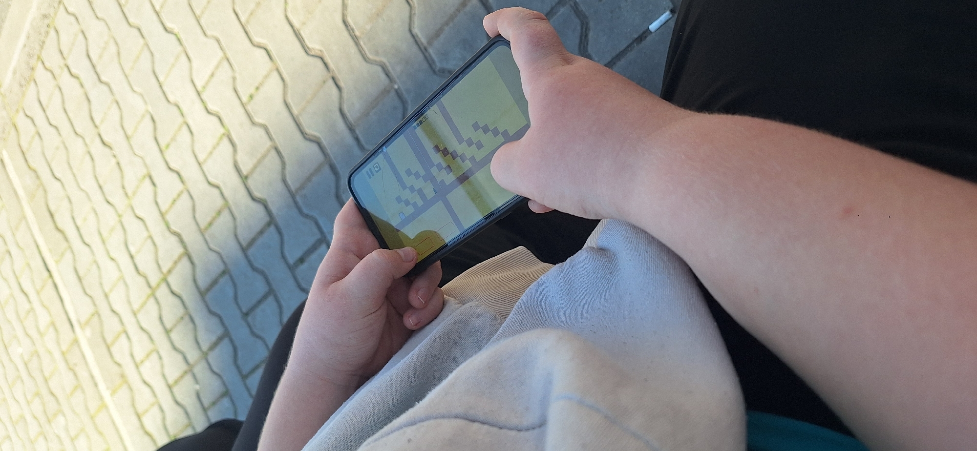 A person is holding a smartphone, playing a pixelated game. The smartphone is held horizontally. The person is sitting down, wearing black trousers and a light-colored sweatshirt. The background shows pavement stones. The location is likely Stara Zagora, Bulgaria, as implied by the prompt.