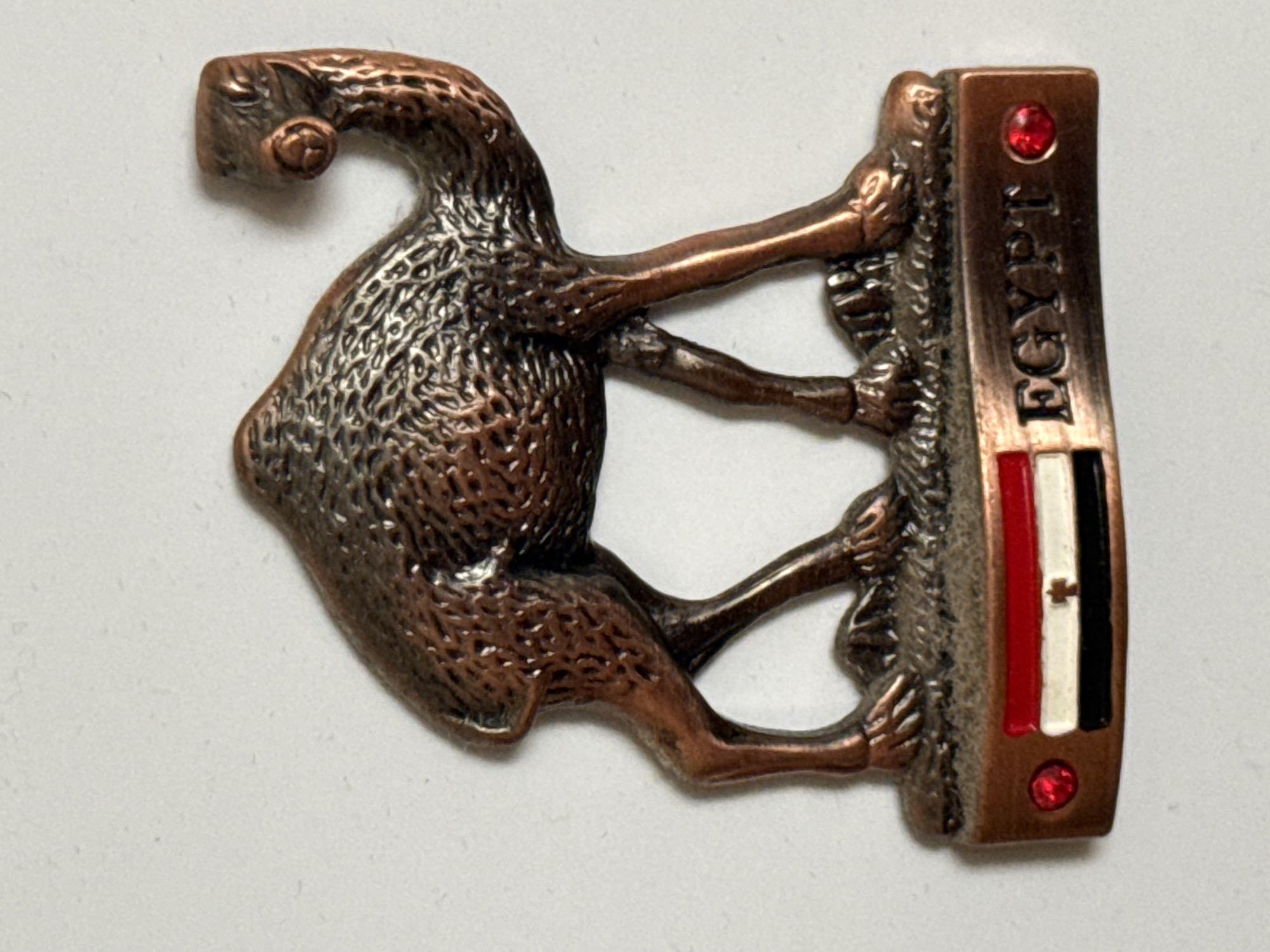 This is a close-up, overhead shot of a copper-colored souvenir or charm shaped like a camel standing on what appears to be a rectangular base. The camel is depicted in a stylized, almost cartoonish manner, with its head bent down and its legs positioned as if it is walking or resting. The body of the camel has a textured surface, resembling rough fur or skin. The camel's legs are slender and seem to be gripping onto the base.
The rectangular base is a darker copper color and has the word "EGYPT" embossed vertically on its side. Below the embossed text, the Egyptian flag is depicted as three horizontal stripes: red on top, white in the middle, and black on the bottom. The white stripe features a subtle emblem, possibly the Eagle of Saladin. At the top and bottom of the base, there are small, red, faceted stones that resemble gemstones.
The background is a plain, light-colored surface, likely a table or a similar flat area, which provides a neutral backdrop that highlights the metallic sheen and details of the object. The lighting is even, with no strong shadows, suggesting it might be taken with a flash or in a well-lit environment. The focus is sharp on the object, with the background slightly softer. The overall impression is that of a small, decorative item.