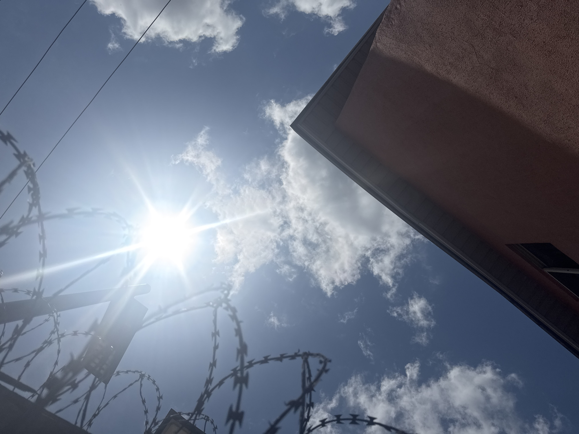 The image is an upward shot taken outdoors, likely in Ihiagwa, Nigeria, given the prompt. The scene includes a bright sun obscured by some haze and a few clouds, with a portion of a building in the upper right corner, presenting a textured, reddish-brown surface. A fence with barbed wire is visible in the lower left quadrant, creating a strong foreground element against the bright sky. There are electrical wires stretching across the upper left area of the sky. There are no people visible.