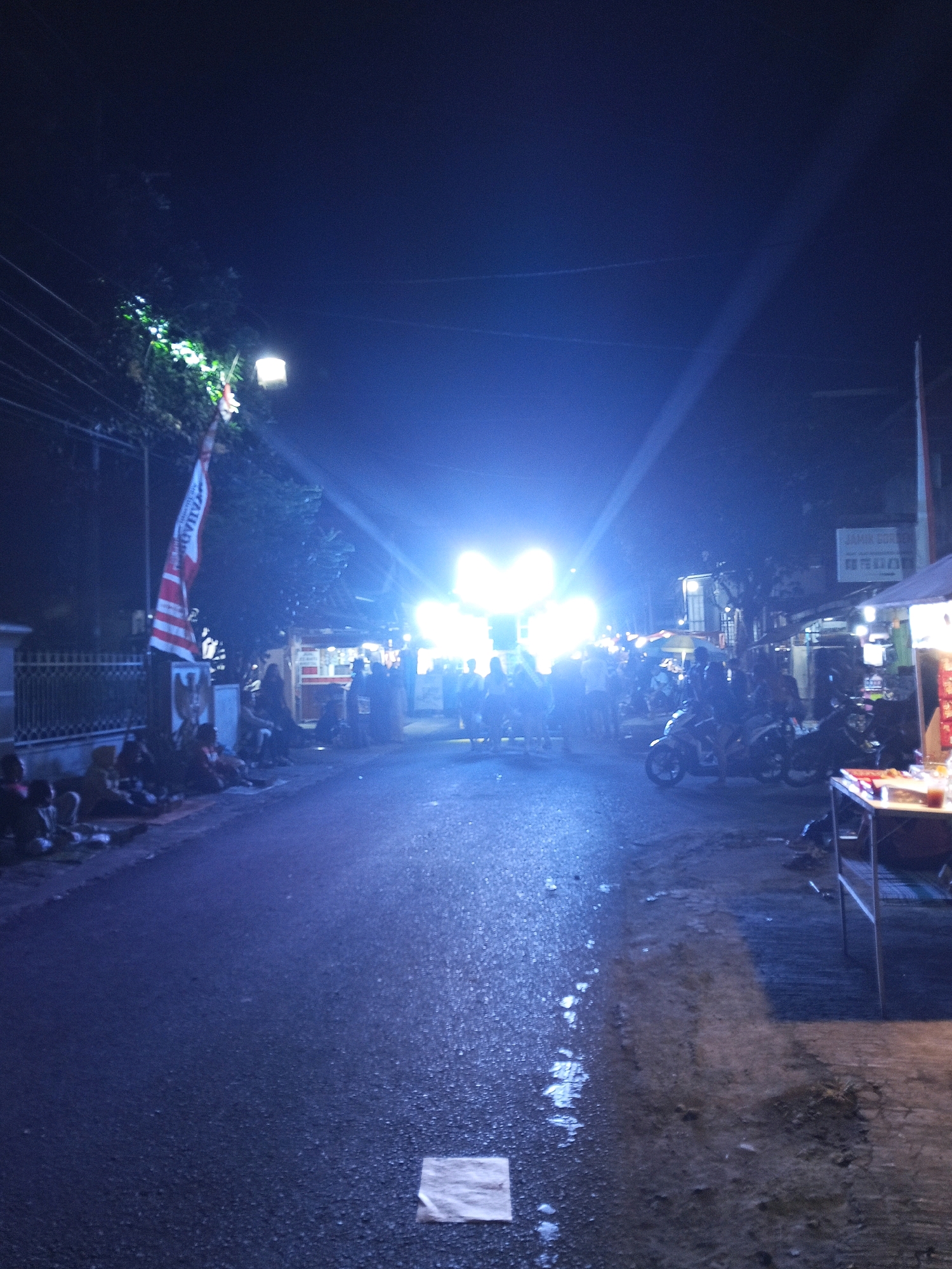 This is a nighttime street scene in Bendosewu, Indonesia, illuminated by bright artificial lights from a stage or event setup. The road appears wet, reflecting the intense lights, and there are several people gathered along the side of the road, some sitting or standing. A flag with red and white stripes is visible, attached to a pole. Several motorcycles are parked or present on the right side of the road. A stall or vendor setup with a table is also on the right. The overall atmosphere suggests a public gathering or event taking place at night.