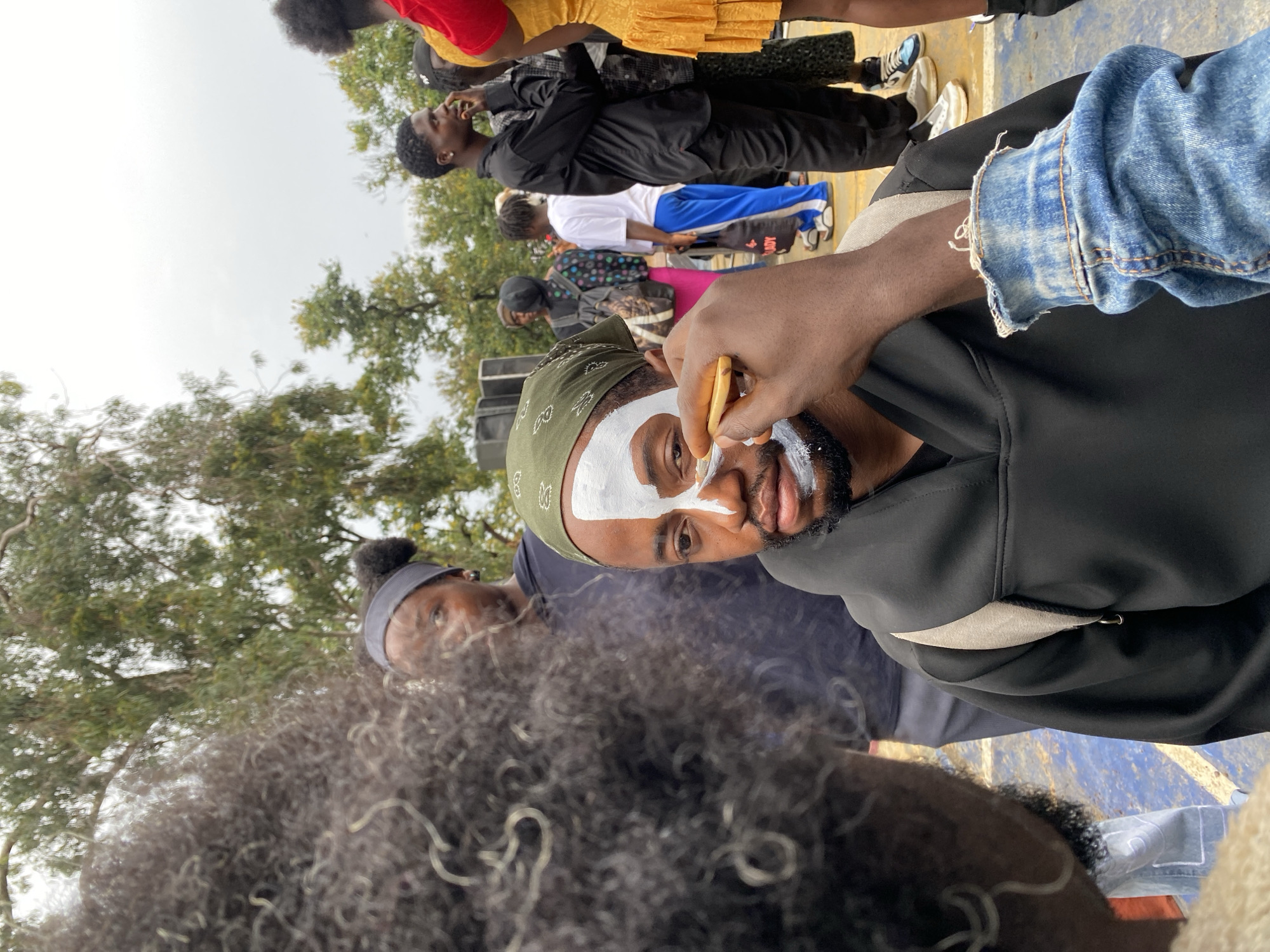 In Bar Jirgi Summa, Nigeria, a man with dark skin, wearing a green bandana and a black hooded garment, has white face paint applied to the left side of his face, covering his eye and cheekbone. A person's hand, wearing a blue denim jacket with frayed cuffs, is holding a thin brush and applying the paint to the man's face. The man is looking towards the person applying the paint. Behind them, other individuals are visible, including a woman with a dark headband and a woman in the foreground with dark, curly hair. Trees and an overcast sky form the background. The scene appears to be outdoors, possibly at an event or gathering.