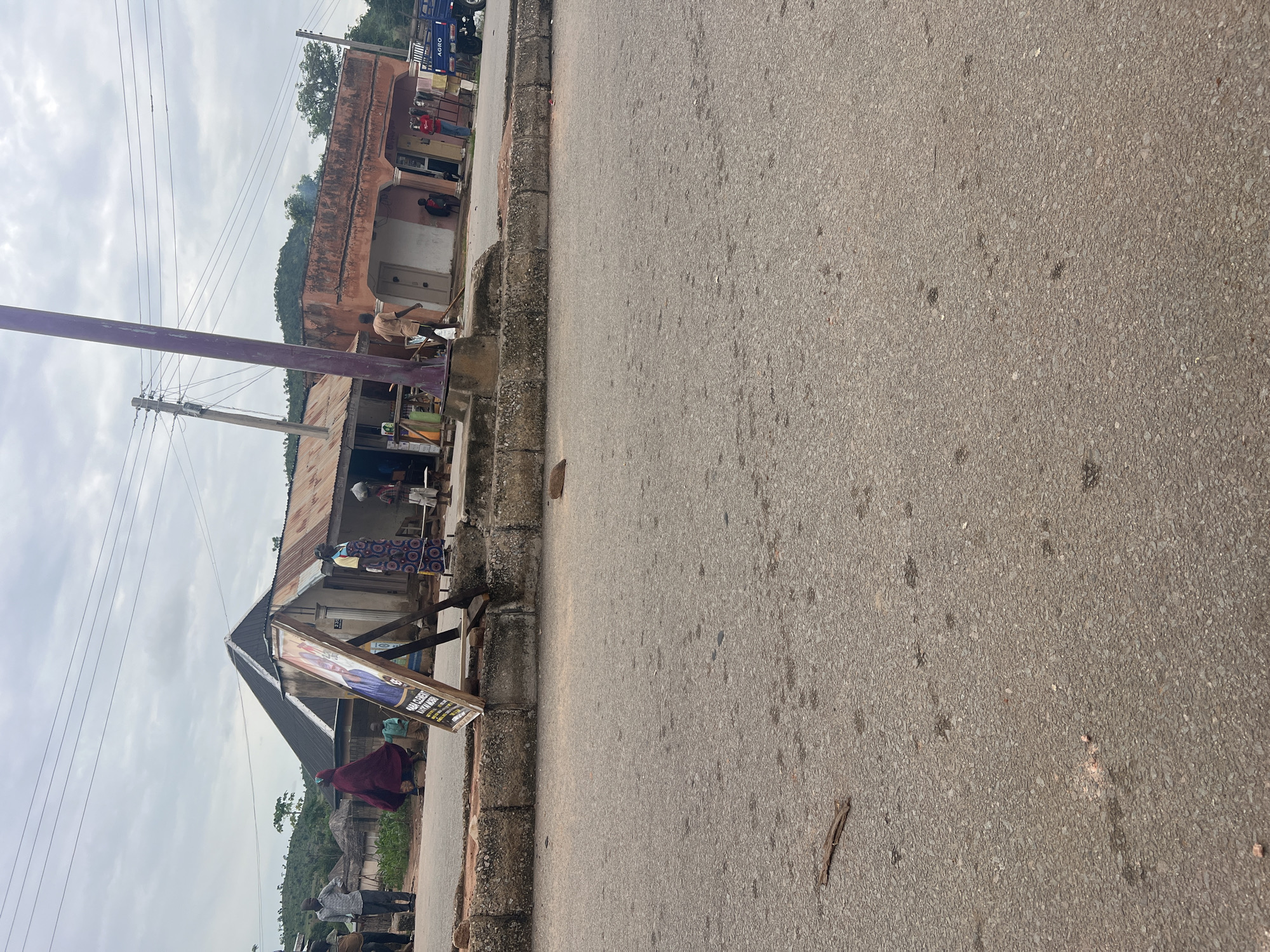 In Kona Garu, Nigeria, a street scene unfolds under an overcast sky. A dirt road runs alongside a concrete barrier. In the background, several structures are visible, including a building with a corrugated metal roof and an orange-colored building with arched doorways. A purple lamppost stands prominently in the foreground.
Several people are present. On the left, a man in a white shirt and dark pants stands near a sign. To his left, another man in a brown shirt and dark pants is visible. A woman in a burgundy garment walks by a child in a light-colored outfit.
In front of the corrugated metal building, a wooden easel holds a sign with a picture of a person and text. A woman in a colorful patterned dress stands near a young child. Further to the right, a man in a brown shirt stands outside the orange building, near another person in red. A blue truck with "AGRO" written on its side is parked to the far right. The ground is paved with asphalt and has some debris.