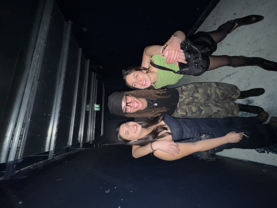 Three young adult females are depicted posing together in a dimly lit, narrow indoor corridor or hallway. The walls are dark, possibly black, with some visible scuffs. The ceiling features exposed metallic channels or tracks, and a green illuminated exit sign, consistent with European standards, is visible in the upper background. The floor is a light, possibly concrete, surface.
From left to right:
The first individual has long dark hair and wears a dark denim strapless top or dress, along with a small black shoulder bag. Her right hand holds a lit cigarette or vape, resting against her hip. She smiles broadly at the camera.
The central individual wears a black beanie covering part of her long dark dreadlocks or braids, clear-framed glasses, a black t-shirt, camouflage cargo pants, and dark boots. She has a silver necklace and smiles broadly. Her left arm is around the shoulder of the person to her right.
The third individual, with dark hair pulled back, wears a light green top, black shorts, sheer black tights, and dark cowboy boots. She carries a black, studded shoulder bag. Her left arm is around the waist of the central individual, and she smiles widely with her eyes partially closed.
All three subjects are smiling directly at the camera, indicating a posed photograph, and their physical contact suggests a friendly interaction. The exact venue within Paris, France, cannot be determined from the visual evidence, but the environment, including the type of exit sign, is consistent with an indoor public space in a European city.