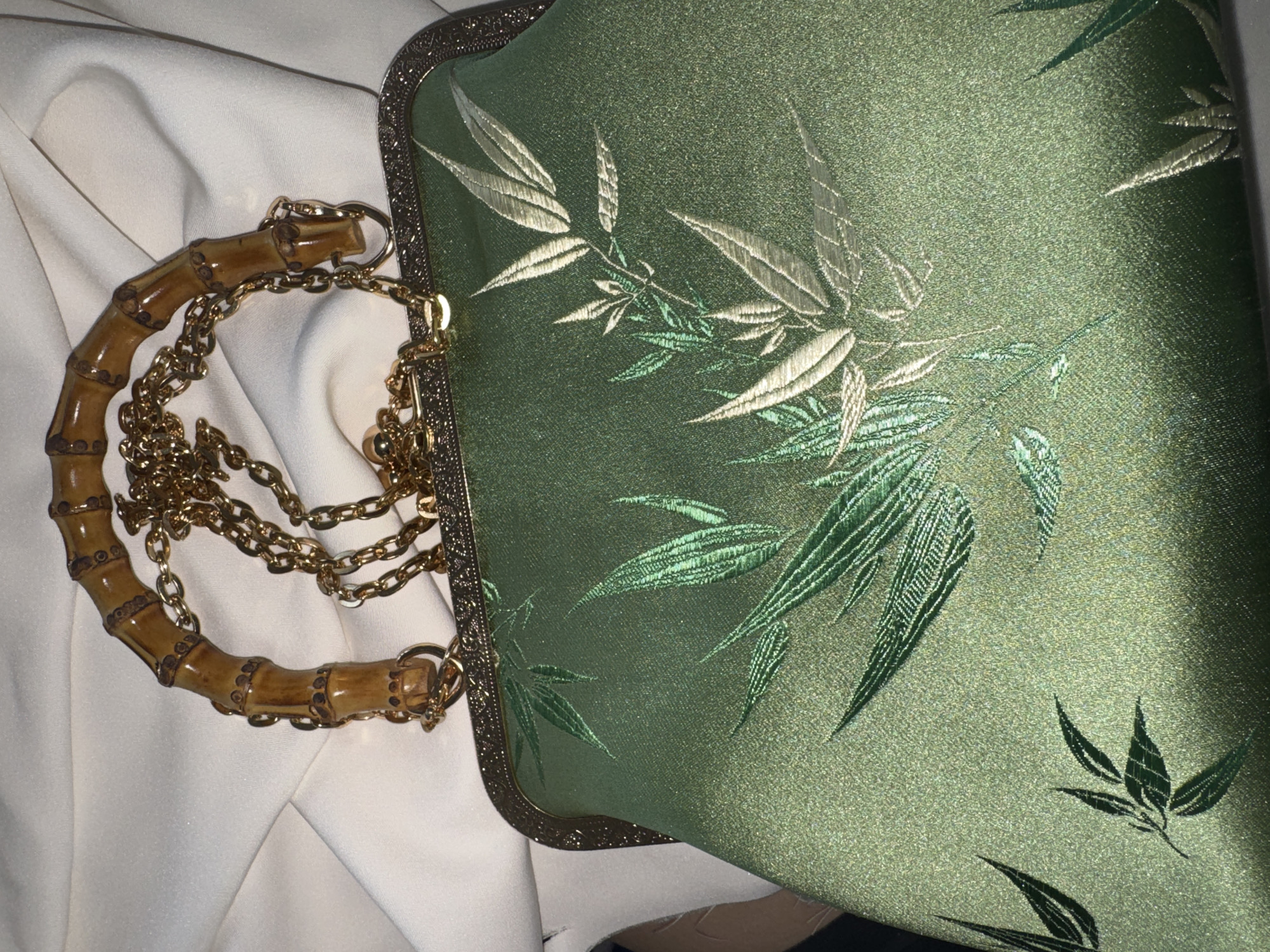 A green, embroidered clutch purse with a bamboo handle and gold chain is displayed on a white surface. The purse features an embroidered bamboo leaf pattern in shades of green and gold. The bamboo handle is curved and segmented, and it connects to the purse with gold chain links. The white surface appears to be fabric, possibly clothing or a tablecloth. The lighting is dim, casting a shadow on the left side of the image.
