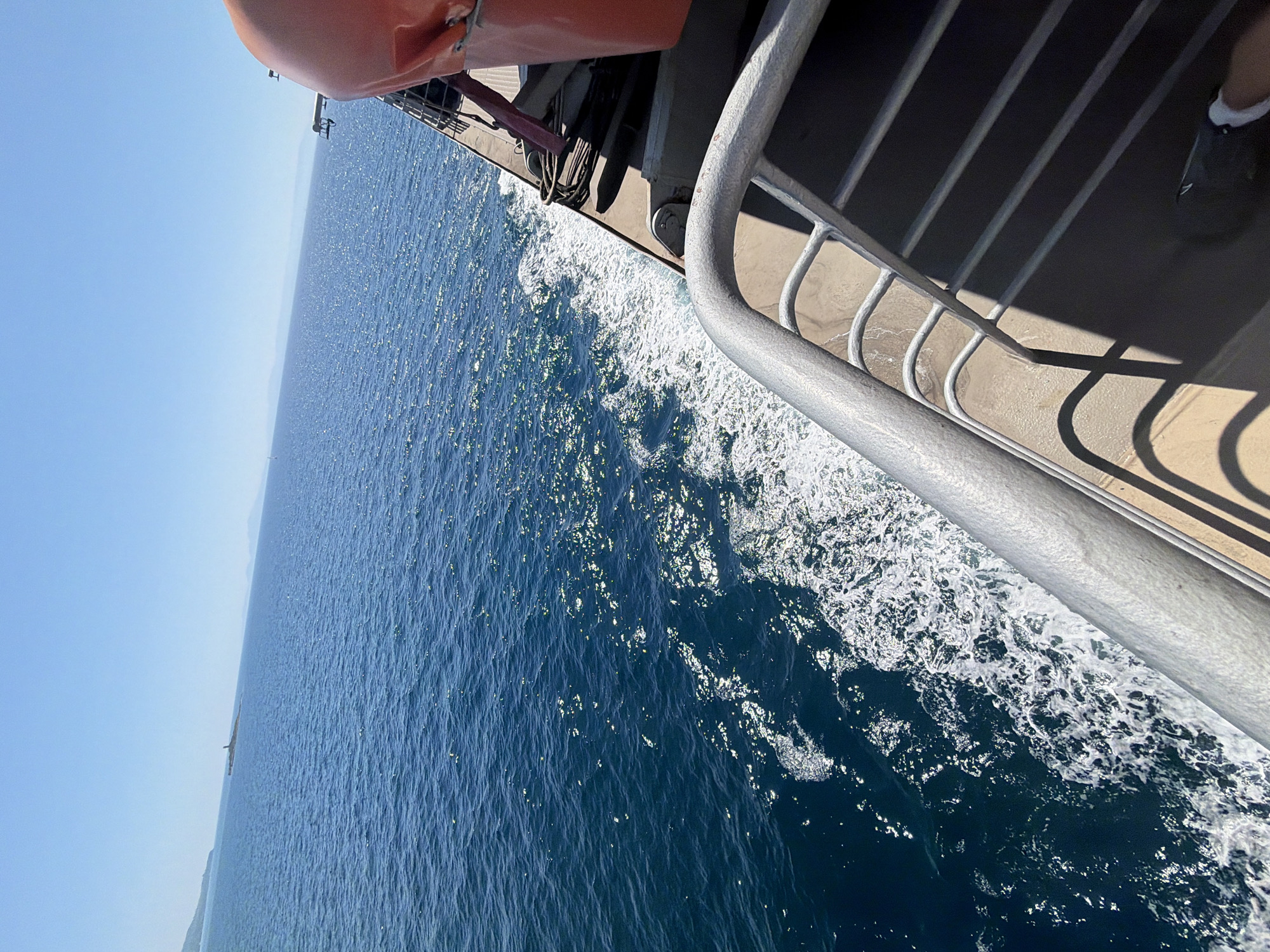 An exterior, high-angle, medium shot captures a bright, sunny day over the ocean from the deck of a moving vessel, likely a ferry, in the Municipal Unit of Kastro - Kyllini, Greece. The foreground shows a metal railing on the deck of the vessel, casting long shadows on the tan-colored deck. A life preserver, colored bright orange, is visible to the upper right. The middle ground is dominated by the deep blue sea, with white wakes trailing from the vessel. The horizon line is clear, with distant landmasses visible, including a small island with a lighthouse-like structure. The sky is a clear, bright blue with no clouds.