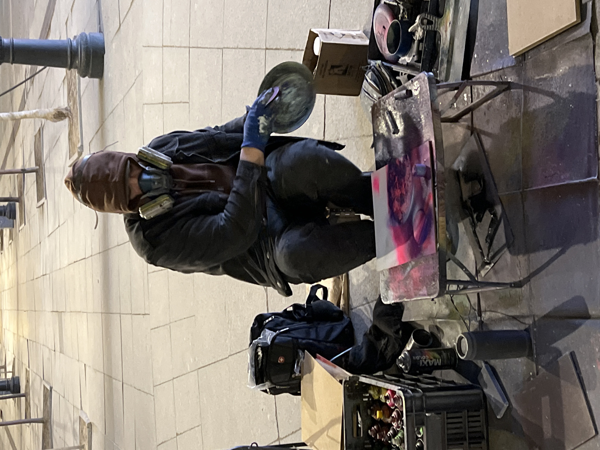 A street artist, equipped with a respirator and gloves, is shown in the process of creating art in Saint Petersburg, Russia. The individual is dressed in dark clothing and is kneeling next to a makeshift workstation composed of a metal table and a crate overflowing with spray paint cans. On the table, a piece of artwork is visible, showcasing an abstract mix of pink and purple hues. The artist holds a round object in their gloved hands, presumably used in the artistic process. The environment consists of stone pavement, a backpack, and a lamppost, which are typical features of the city's urban landscape.