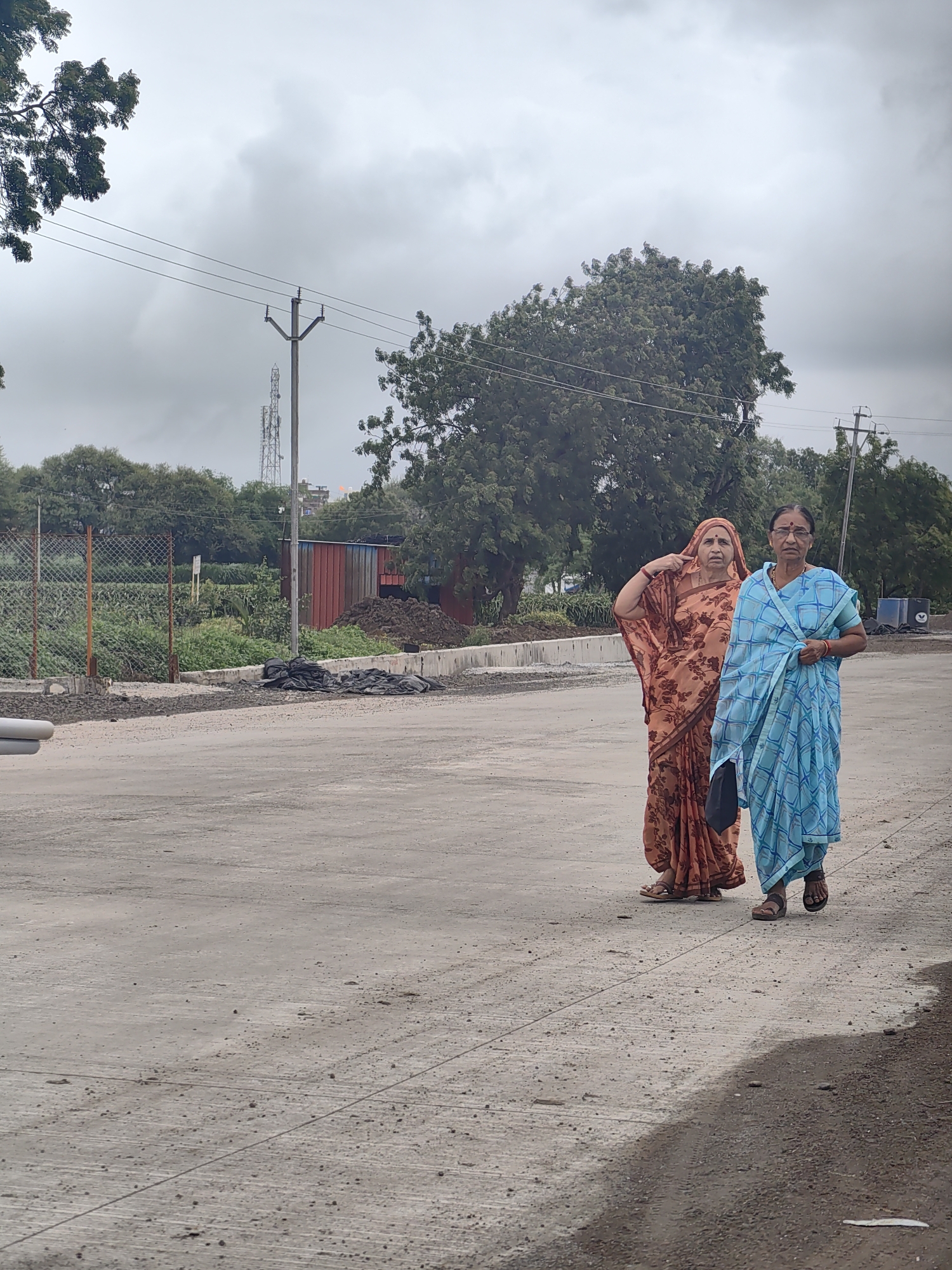 Two women, one in an orange patterned sari and the other in a blue patterned sari, are walking on a paved surface in Walsawangi, India. The woman in orange is gesturing towards her head. The background includes trees, a chain-link fence, a building with a red corrugated metal side, a pile of dark earth, and power lines against an overcast sky.