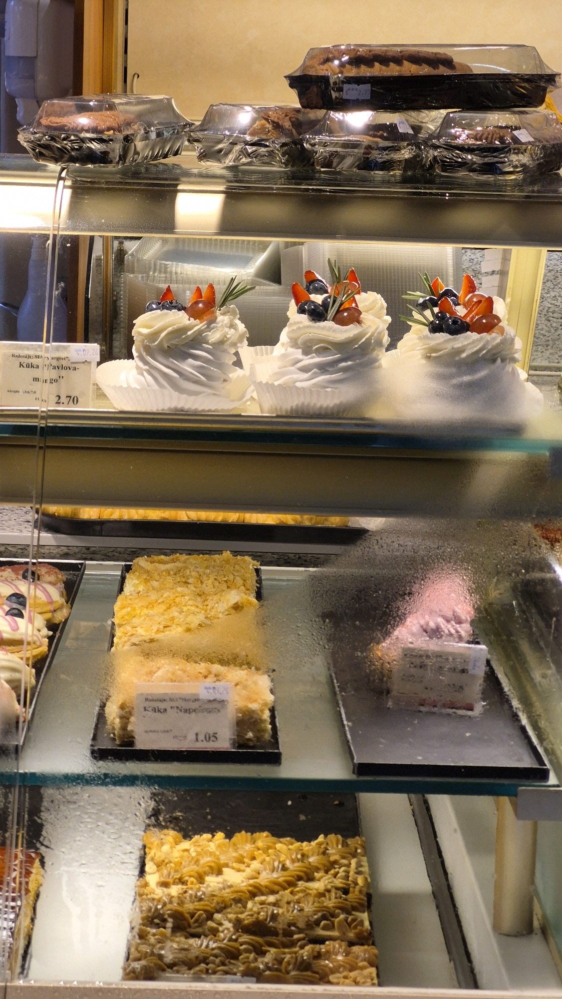 The image displays a refrigerated glass display case filled with various desserts. On the top shelf, there are several pre-packaged baked goods in clear plastic containers. The middle shelf features three individual meringue-based desserts topped with whipped cream, fresh berries (strawberries and blueberries), and garnishes. A price tag indicating "2.70" is visible for one of these desserts. Below this, another shelf holds a rectangular cake with a flaky, golden-brown topping, and a price tag displaying "1.05" and the text "Kūka "Napoleons"". To the right of this cake, another dessert is partially visible, also with a price tag. The bottom shelf exhibits a large rectangular dessert with a textured, caramel-colored topping. The environment appears to be a bakery or patisserie, with warm lighting and condensation on the glass, suggesting a cold interior. The location is identified as Jēkabpils, Latvia.
