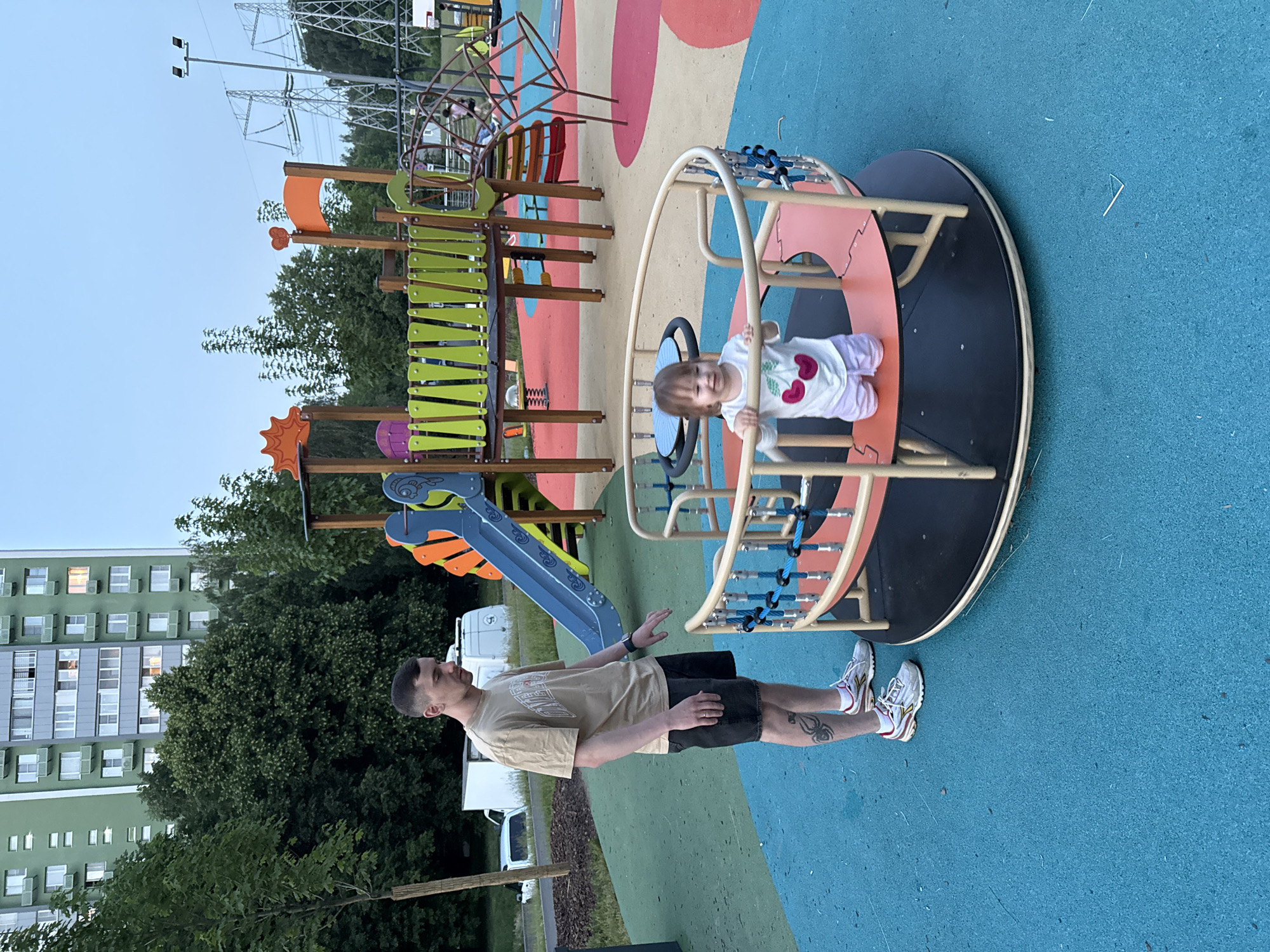 In Moscow, Russia, a young man stands on a bright blue playground surface next to a merry-go-round. He is wearing a beige t-shirt, dark shorts, and white sneakers, with a tattoo on his left leg. A toddler in a white t-shirt with cherry prints and light purple shorts is on the merry-go-round, holding onto its railing. In the background, a colorful playground structure with slides and climbing elements is visible. Beyond the playground, a tall green apartment building and trees can be seen, with power lines and towers in the distance, under a pale sky.