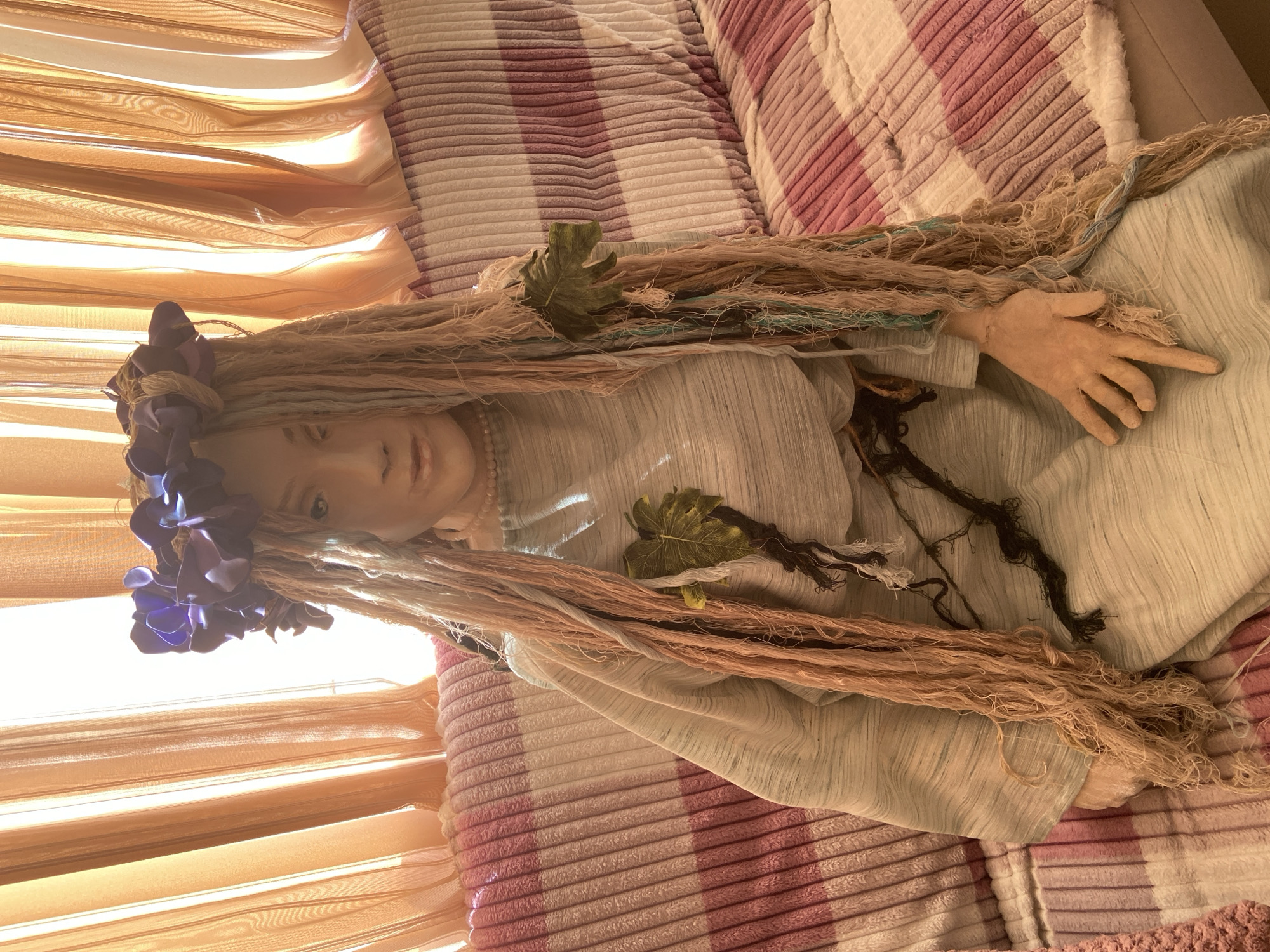 The image shows a doll lying on a striped blanket. The doll has a floral crown, fair skin, and long, fibrous hair. Some green leaves and dark fibers are interspersed in the doll's hair. The doll is wearing a light-colored garment. Background includes vertical window blinds. Location is Chaikovsky, Russia.