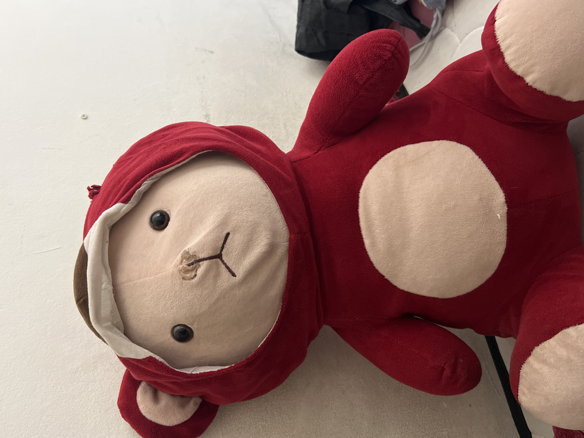 A stuffed toy bear, predominantly red with a tan face and white oval on its chest, is positioned leaning against a white wall. The bear is wearing a red hooded costume with brown and white trim around the face opening. Its eyes are black, and its nose and mouth are embroidered in black thread. The background shows a portion of a white wall with a small, circular mark, and a hint of other objects to the right, including what appears to be a black bag. The image is taken from a slightly low angle, focusing on the bear's head and upper body. There is no indication of its location in Denpasar, Indonesia.