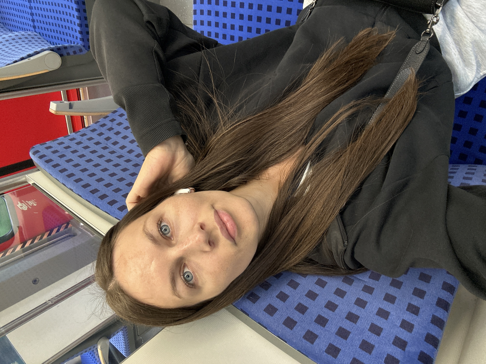 A young woman with long, straight brown hair and clear blue eyes is shown lying across two blue, patterned train seats. She wears a dark, possibly black, hoodie or sweatshirt, with a white earbud visible in her left ear. Her head rests on one seat, and her body extends onto another, suggesting a relaxed posture during travel. She looks directly at the camera with a calm and neutral expression.
The setting is clearly the interior of a train, featuring blue upholstered seats adorned with a repetitive dark blue square pattern. Metal fixtures and a vibrant red surface are visible in the background; the red surface bears partially legible white text, possibly "DB" (Deutsche Bahn), which aligns with the provided location context of Buxtehude, Germany. The lighting is even, indicating an indoor environment, likely during the daytime. The image captures a serene moment of a passenger resting or taking a selfie aboard a German train.