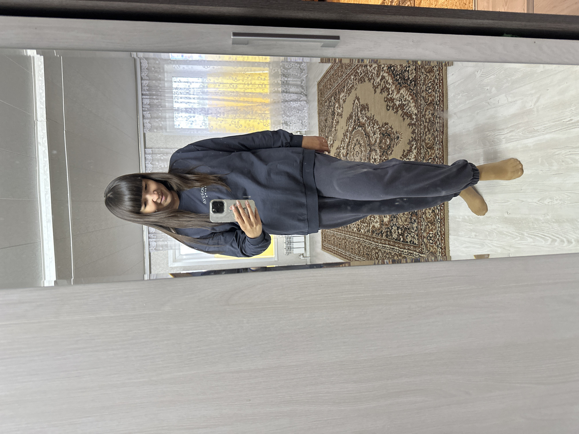 A mirror reflects a woman taking a selfie indoors. The woman is wearing a dark blue tracksuit. Behind her, the reflection shows a room with a window covered by a sheer curtain and a patterned rug on the floor. The location is likely in Среднее Кечево, Russia, based on the available information.