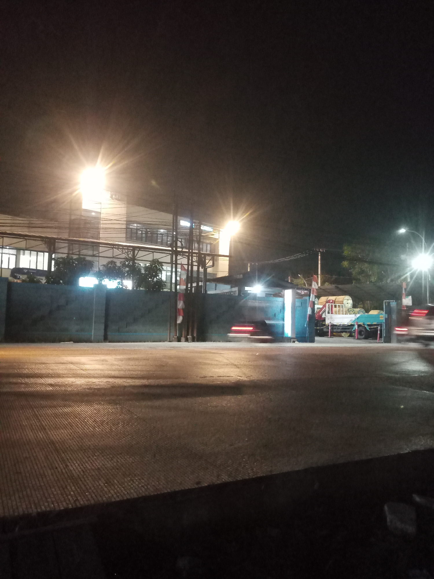 A nighttime, low-light, eye-level shot captures a street scene in Balaraja, Indonesia. The foreground is dominated by a wet, paved road with a ribbed texture, reflecting the artificial lights above. In the midground, a concrete barrier or wall runs horizontally across the frame, separating the road from a building. Behind the wall, a multi-story building with numerous windows is visible, illuminated from within. Several trees are planted in front of the building. The right side of the midground features an open gate or entrance area with several vehicles and structures. A white truck with a crane and a teal-colored trailer are parked. There are also red and white striped poles and a blue structure. Several cars are depicted as streaks of light, indicating movement. The background is dark, with streetlights casting bright, starburst-like glows. Power lines are visible crisscrossing the sky. The overall atmosphere is one of a busy, illuminated urban environment at night.
