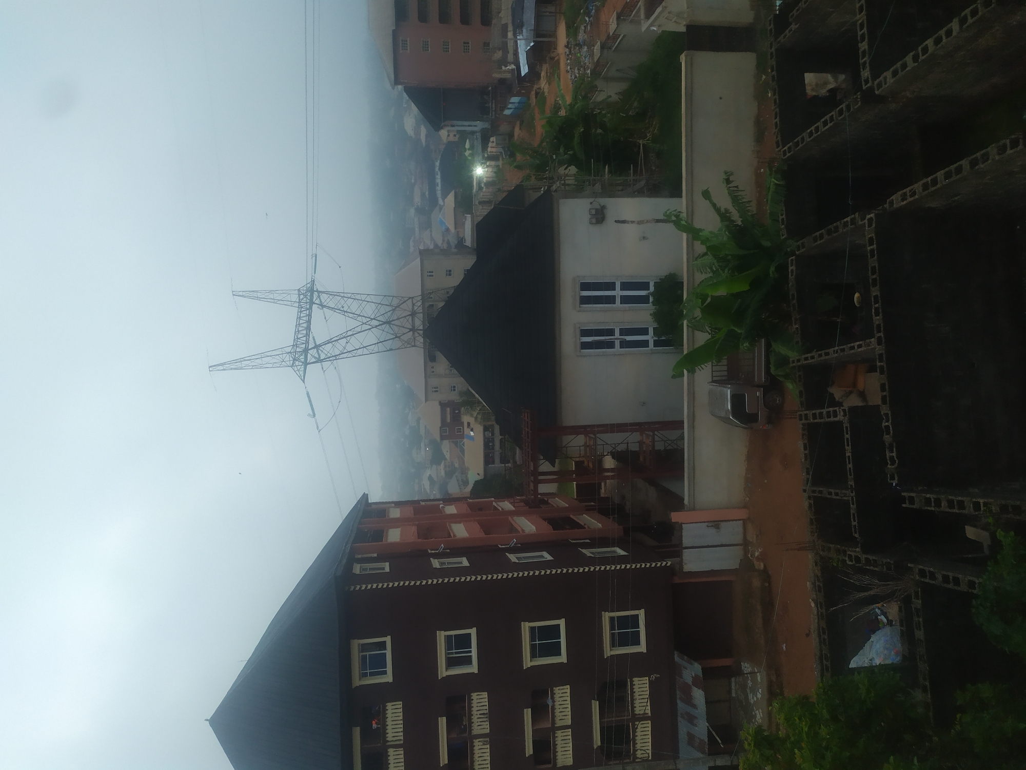 The image depicts a foggy urban scene in an unknown city. A large electrical transmission tower dominates the left side of the frame. Numerous buildings of varying heights and designs are visible. One building is under construction; its skeletal frame is visible on the right side of the picture. A dark, multistory building with light-colored windows is positioned prominently in the foreground. Overhead electrical wires stretch across the sky, adding to the industrial nature of the scene.