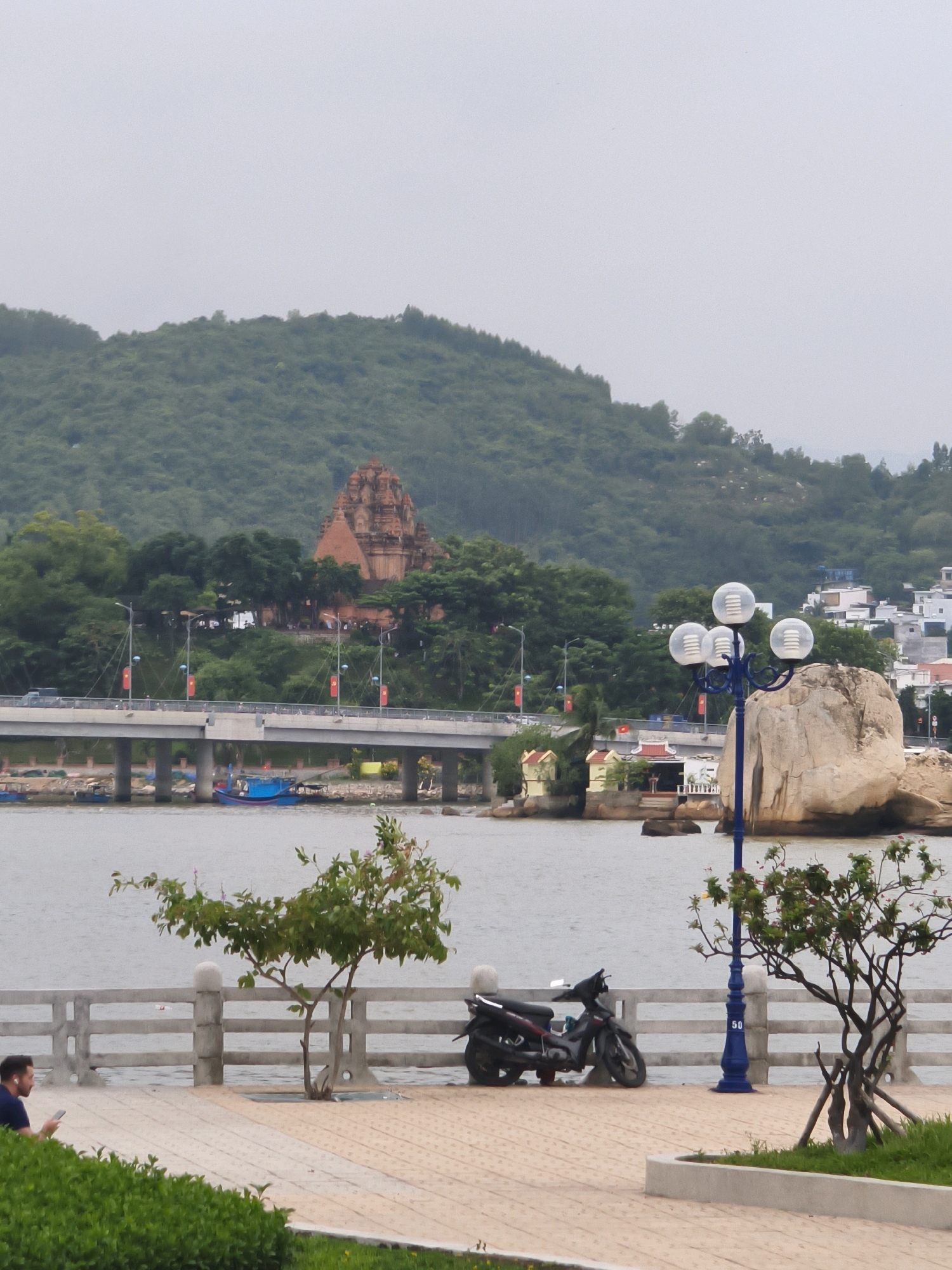 The image depicts a scenic view along a waterfront in Nha Trang, Vietnam. In the foreground, a paved walkway runs parallel to a low concrete barrier overlooking a body of water. A young man is seated on the walkway, looking at his phone. To his right, a small, leafy tree is planted. Further along the walkway, a black motorcycle is parked, and a tall, ornate lamppost with three spherical lights stands next to a large, light-colored rock formation. In the middle ground, a bridge spans the water, with a few small boats visible underneath and on the water. Beyond the bridge, a distinctive red-tiled pagoda-style building sits atop a hill amidst lush green trees. The background is dominated by a verdant, tree-covered mountain under a hazy sky. Buildings are visible on the far right, on the slopes of the mountain. The overall atmosphere is calm and coastal.