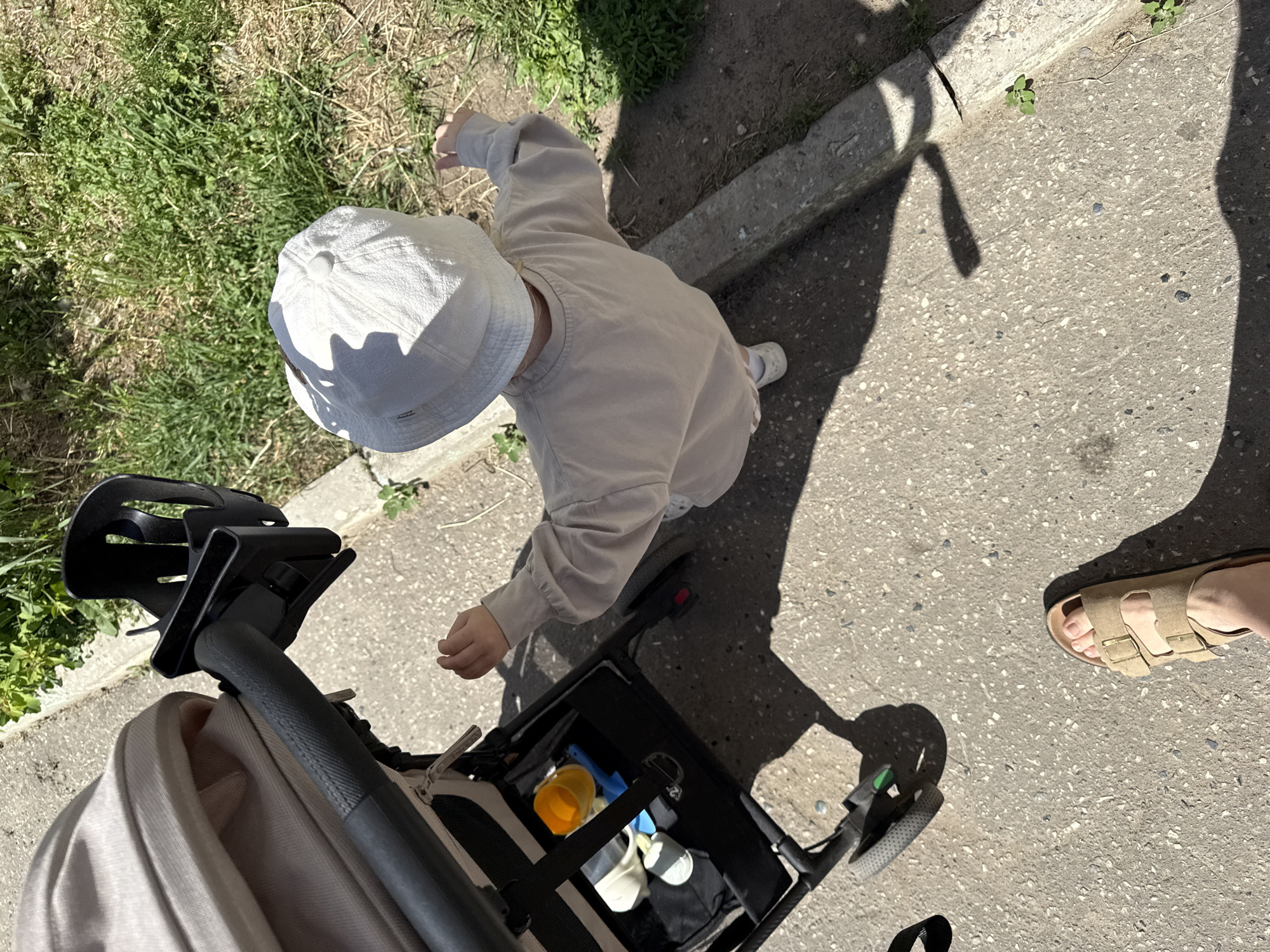 Captured from an overhead perspective, the image portrays a child in a light-colored bucket hat and long-sleeved shirt standing near a stroller on an asphalt surface. The stroller, situated on the lower left, contains toys including a yellow cup and white containers. The child's shadow is cast onto the pavement, alongside the shadow of an individual wearing tan sandals, suggesting the presence of an adult. The setting appears to be near a grassy area adjacent to a concrete curb. The specific location within Vladimir, Russia, cannot be determined from the visual information provided.