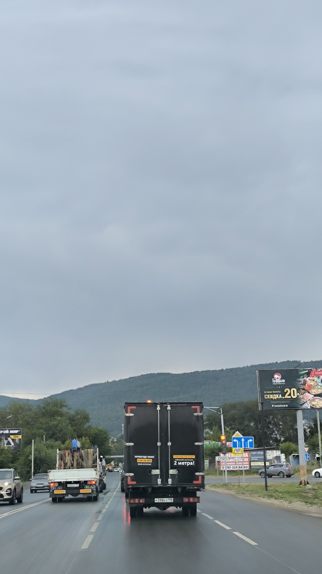 In Miass, Russia, on a wet, overcast day, traffic moves along a multi-lane highway. A black truck with Russian license plates P398KY 196 is in the right lane, displaying the text "2 метра!" on its rear doors. To its left, a white truck carries construction materials. Other vehicles, including a light-colored sedan, are also on the road. Billboards with advertisements are visible on the right side of the highway, and a lush green hillside forms the backdrop. Traffic lights and road signs are present, indicating an urban or suburban setting.